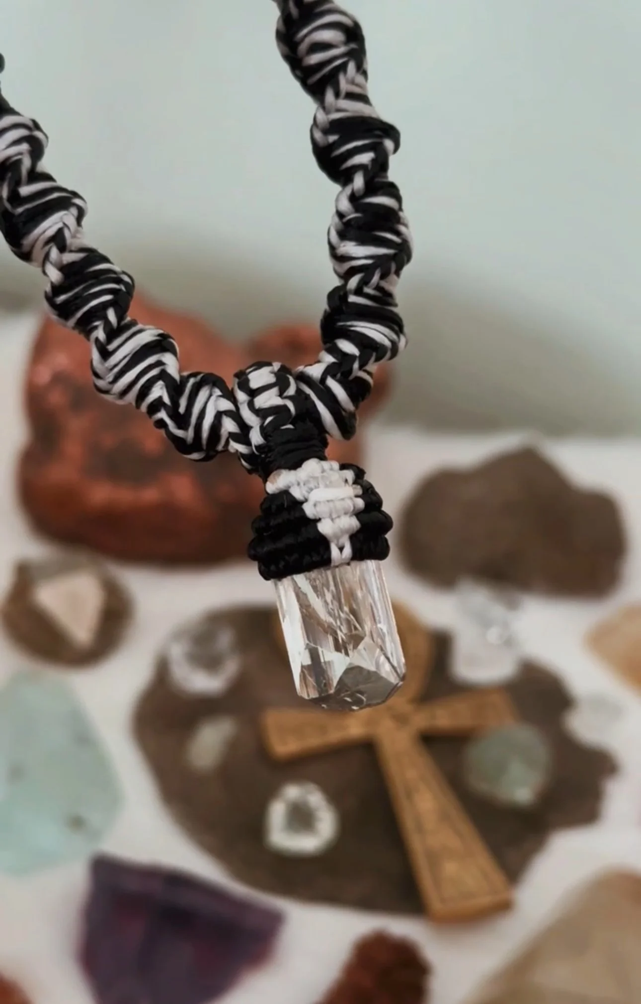 A close-up of a transparent crystal hanging from a black and white twined wrap, with blurred stones and decorative objects in the background.