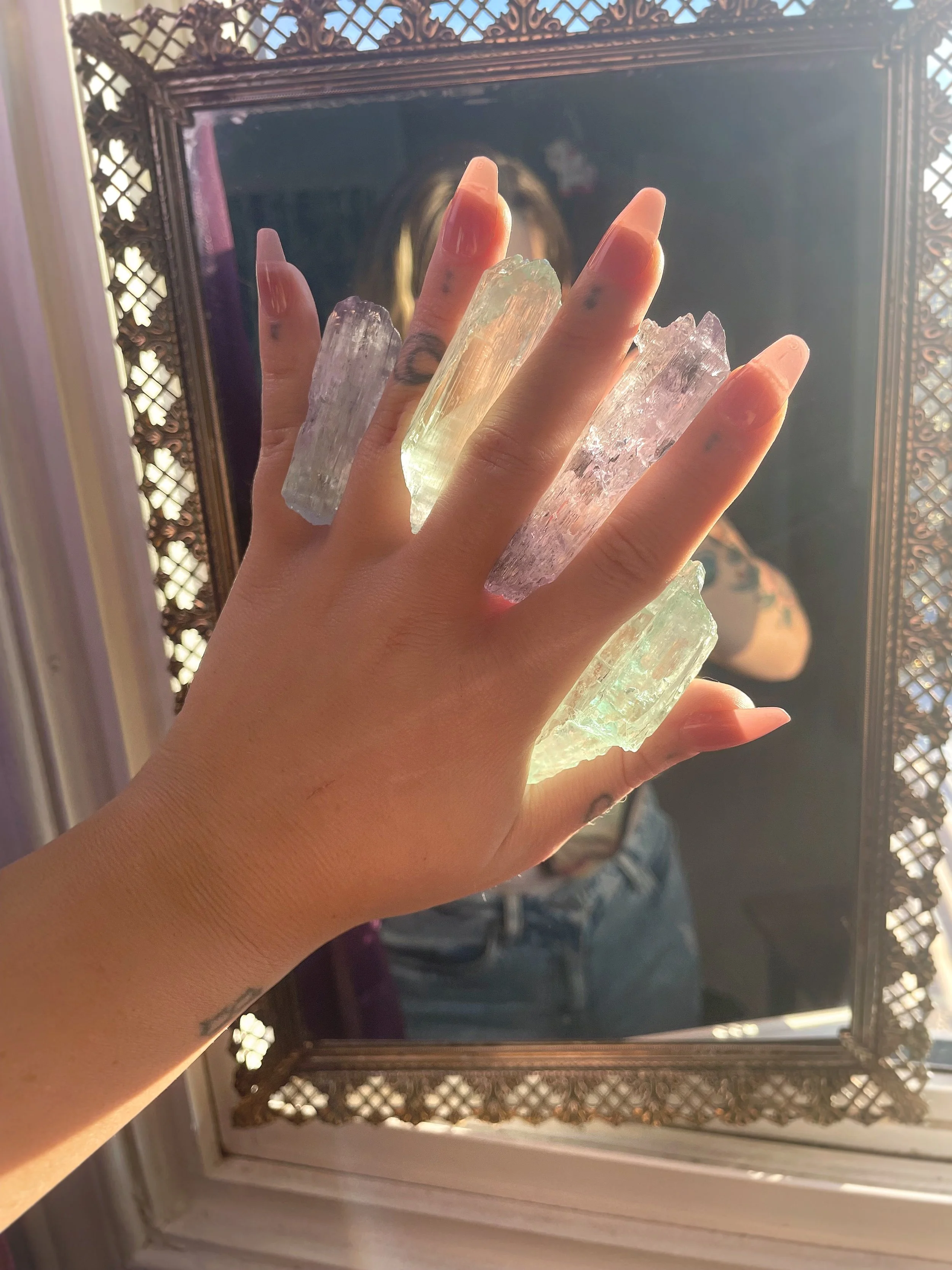A person's hand holding several large, irregularly shaped, translucent crystal stones in front of a mirror with a decorative wooden frame. The person has long, natural nails and some tattoos on the fingers. There is sunlight coming through a window, 