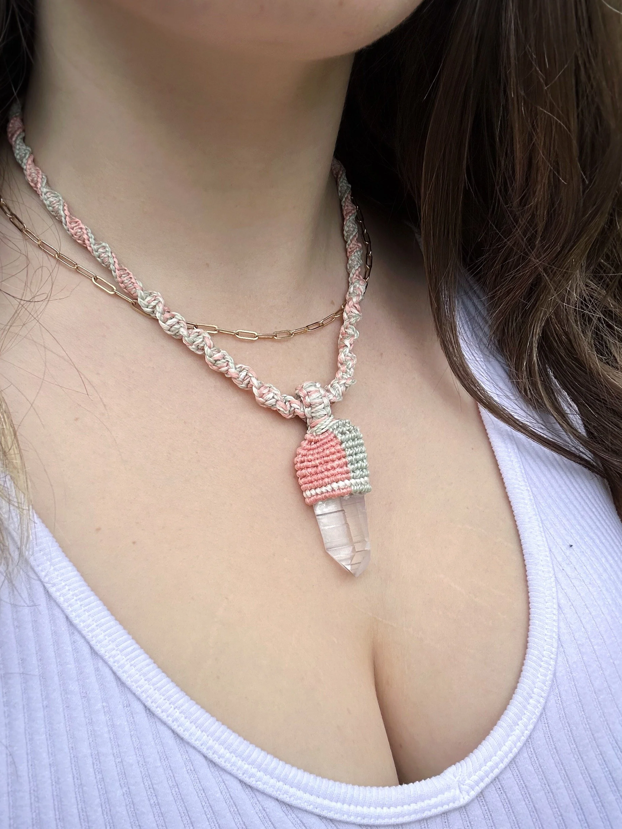 Close-up of a person's chest and neck, with two necklaces: a chain and a macramé necklace with a crystal pendant, wearing a white ribbed V-neck top.