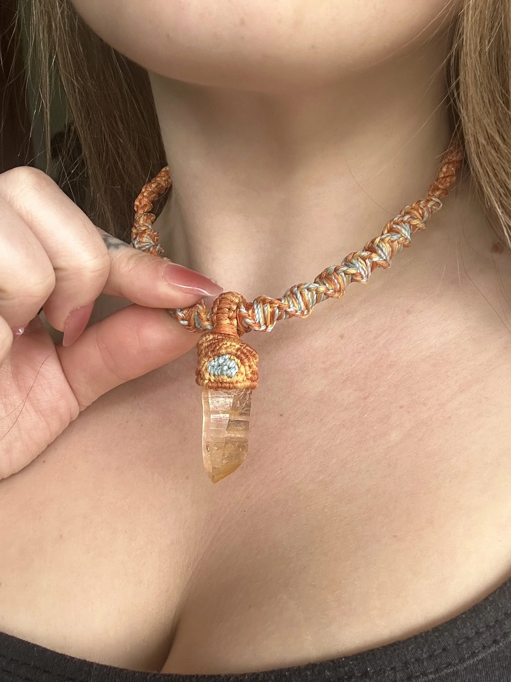 Close-up of a person's neck and upper chest wearing a handmade, knotted necklace with a large, rough, orange gemstone pendant.