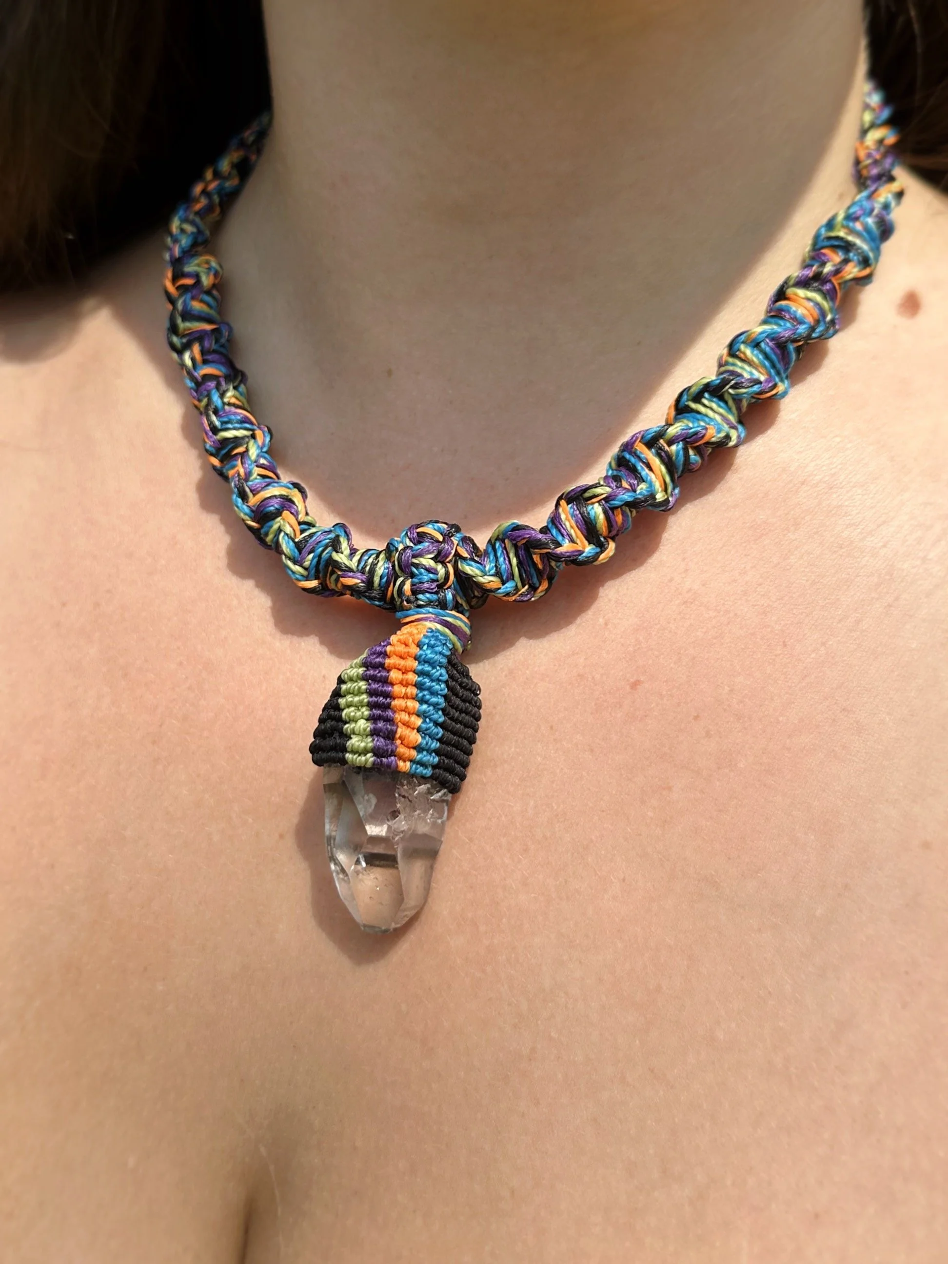 Close-up of a person's neck and chest wearing a colorful, woven necklace with a clear quartz pendant.
