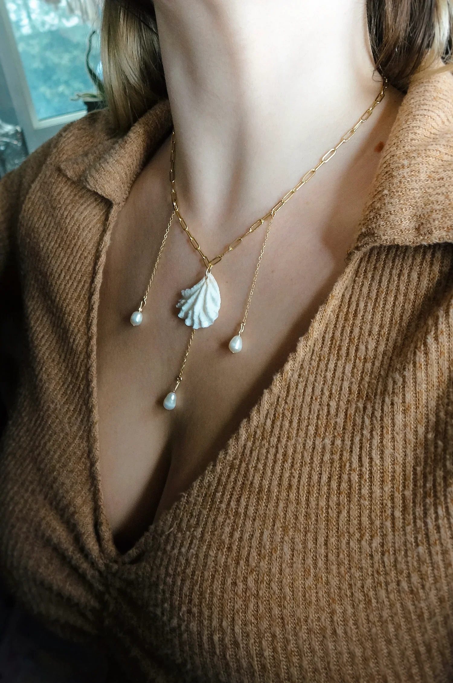 Close-up of a woman's chest wearing layered gold necklaces with pearl and shell pendants, partially covered by a brown ribbed sweater.
