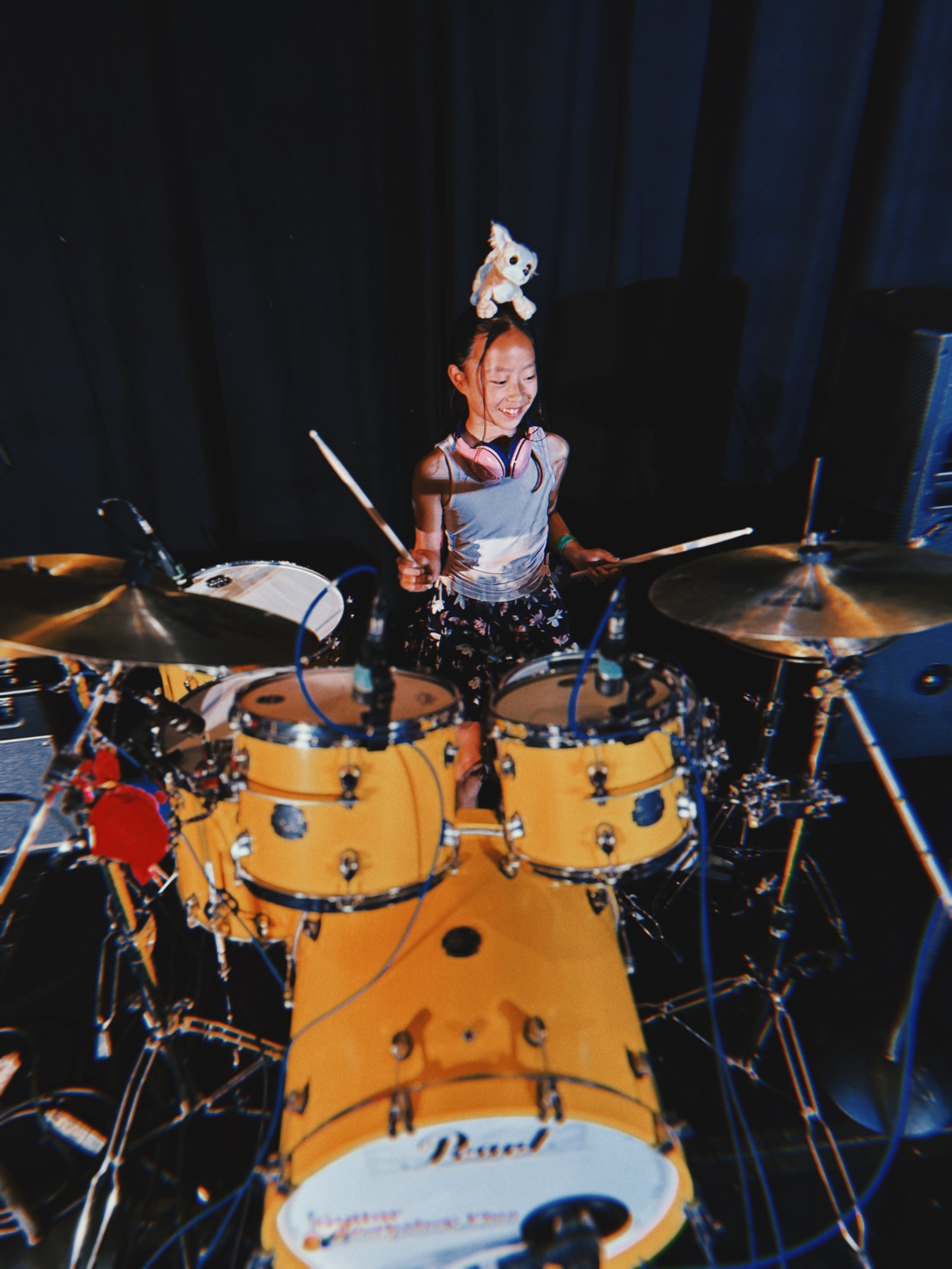 A young girl with a stuffed animal on her head, playing a drum set on stage with a black curtain background.