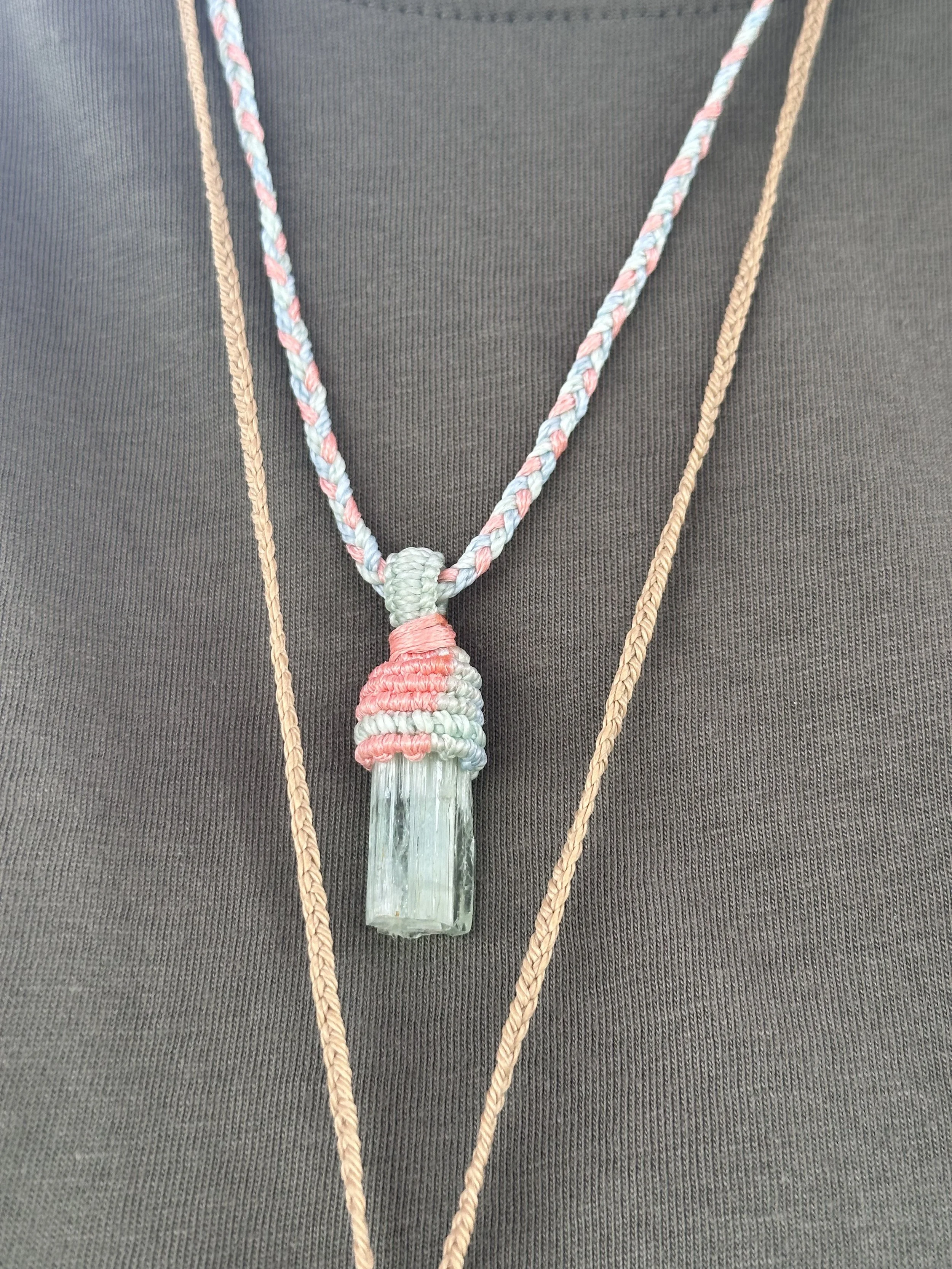 A long, rectangular, clear crystal pendant hanging from a pastel-colored braided necklace, worn over a dark gray shirt, with additional layered necklaces of different lengths around the neck.