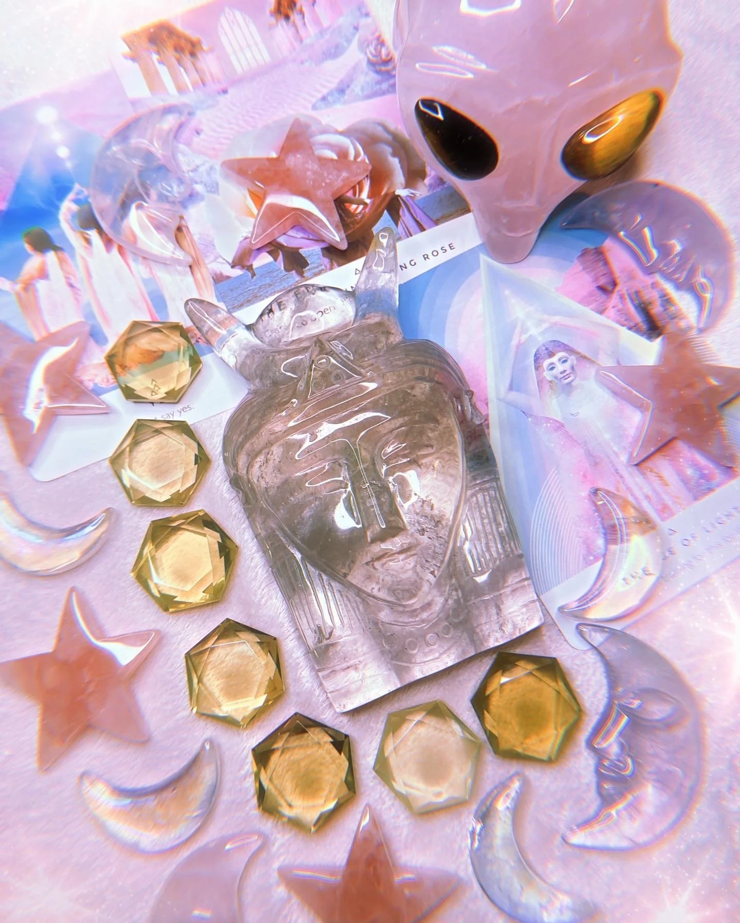 A collection of decorative items including a large pink alien head sculpture with golden eyes, a transparent human face mask, star-shaped and hexagonal gold-colored gems, and magazine pages featuring a woman and a landscape scene with pink and purple