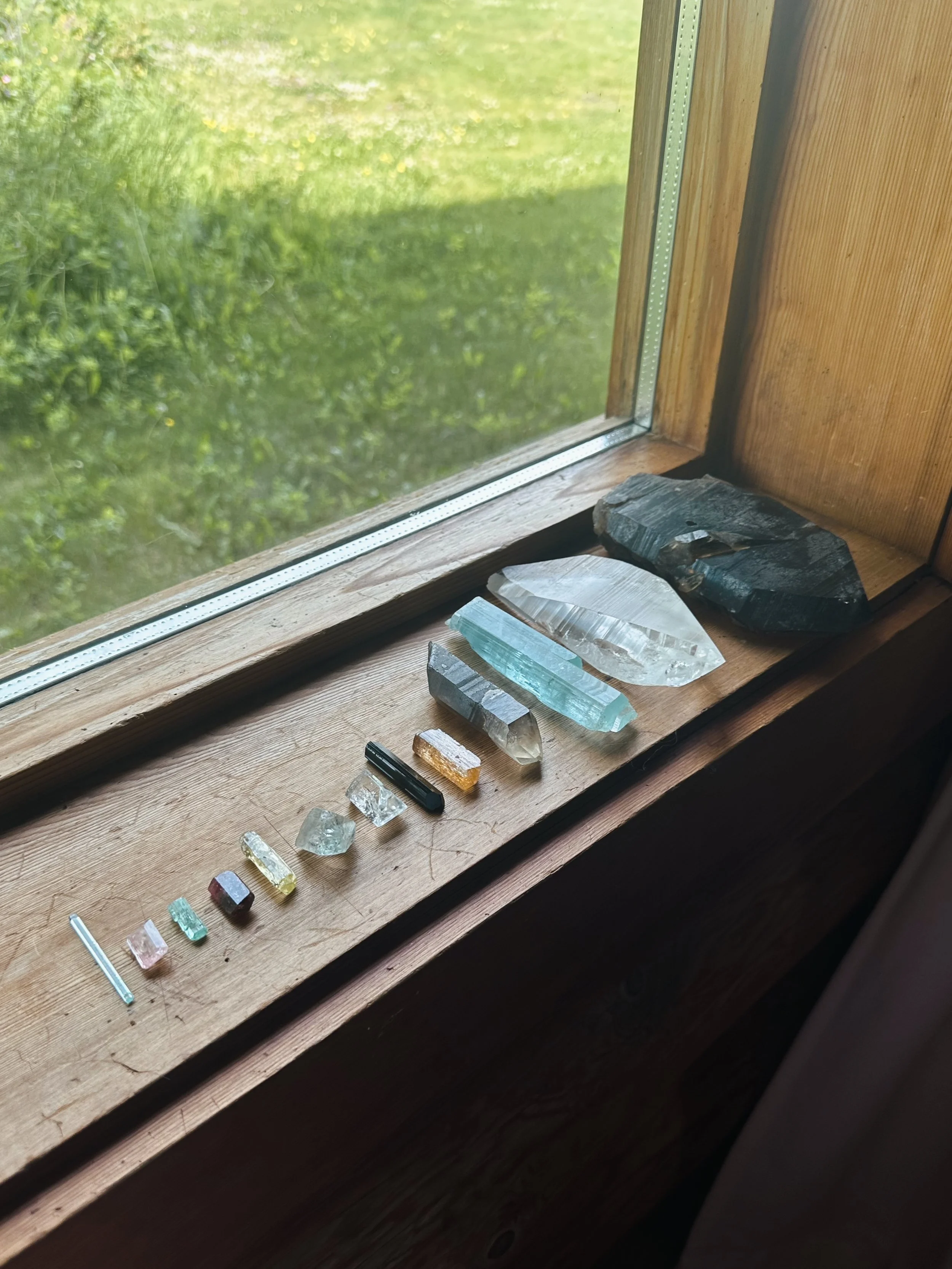 Various colorful crystals and stones arranged on a wooden window sill with a view of a grassy yard outside.