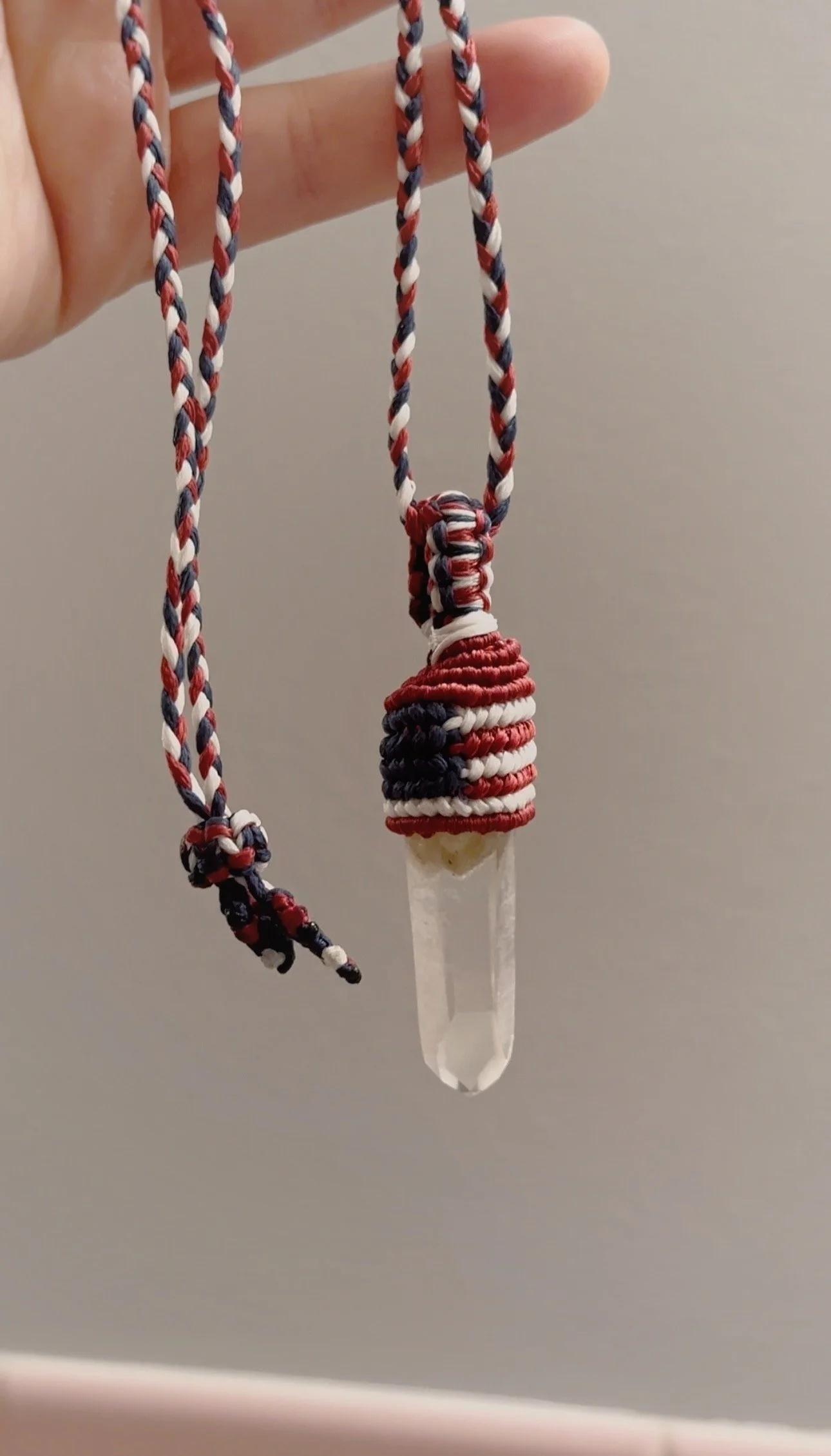 A handmade necklace with a clear crystal pendant wrapped in red, white, and blue embroidery, hanging from a braided cord made of similar colored threads.