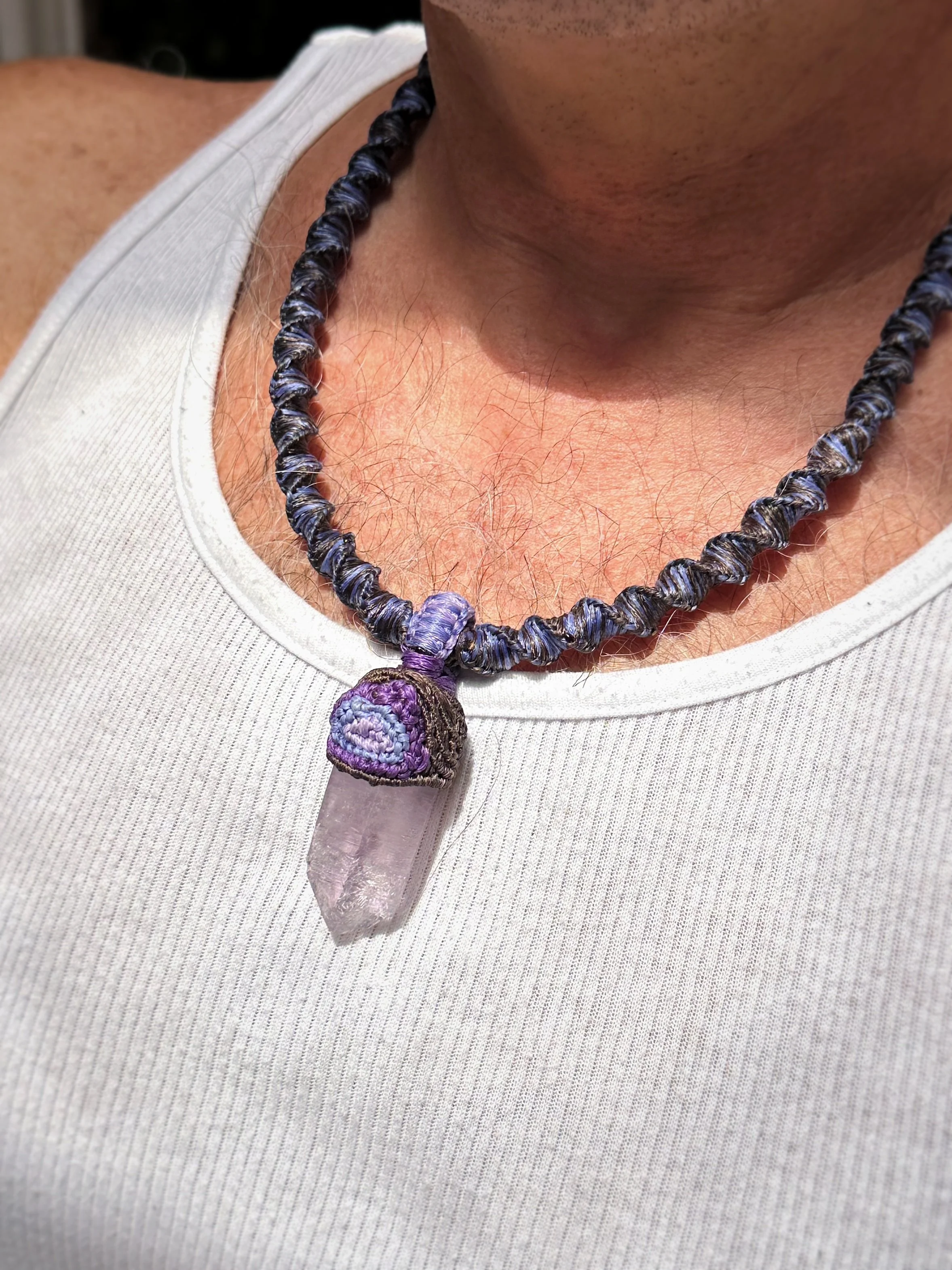 Close-up of a person wearing a necklace with purple, blue, and black beads and a large crystal pendant. The person is wearing a white sleeveless top.