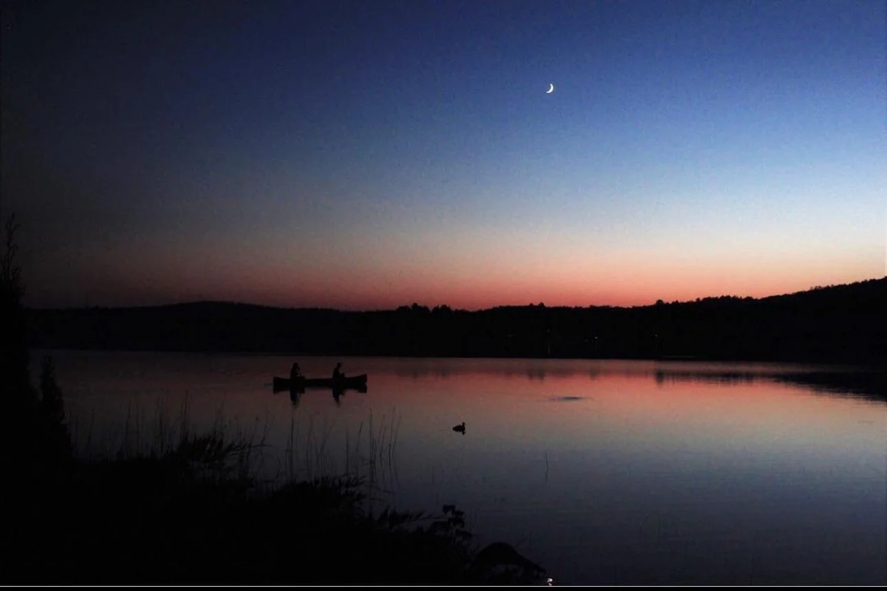 Two people in a canoe on a calm lake during twilight with a crescent moon in the sky.