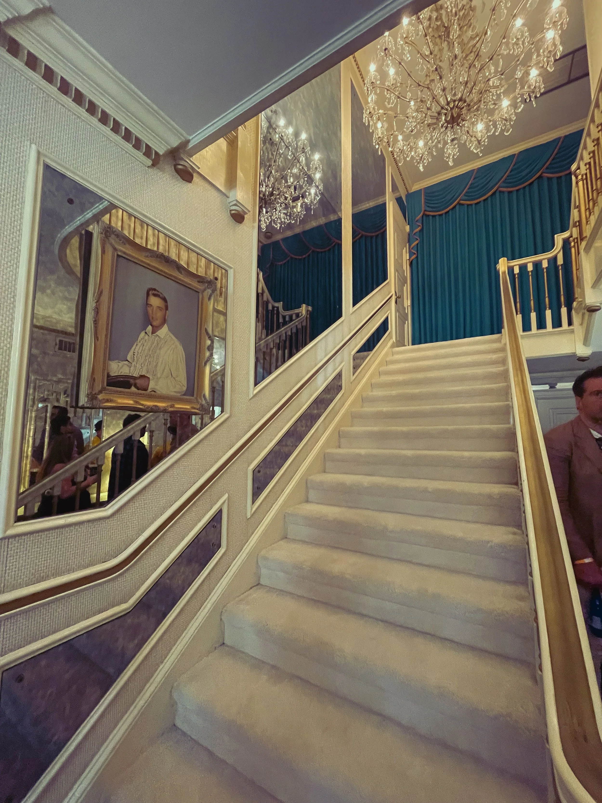An elegant staircase with a white carpet runner, white railings, and a gold and blue curtain at the top. There is a large crystal chandelier hanging from the ceiling and framed artwork on the wall next to the staircase. The overall decor is luxurious