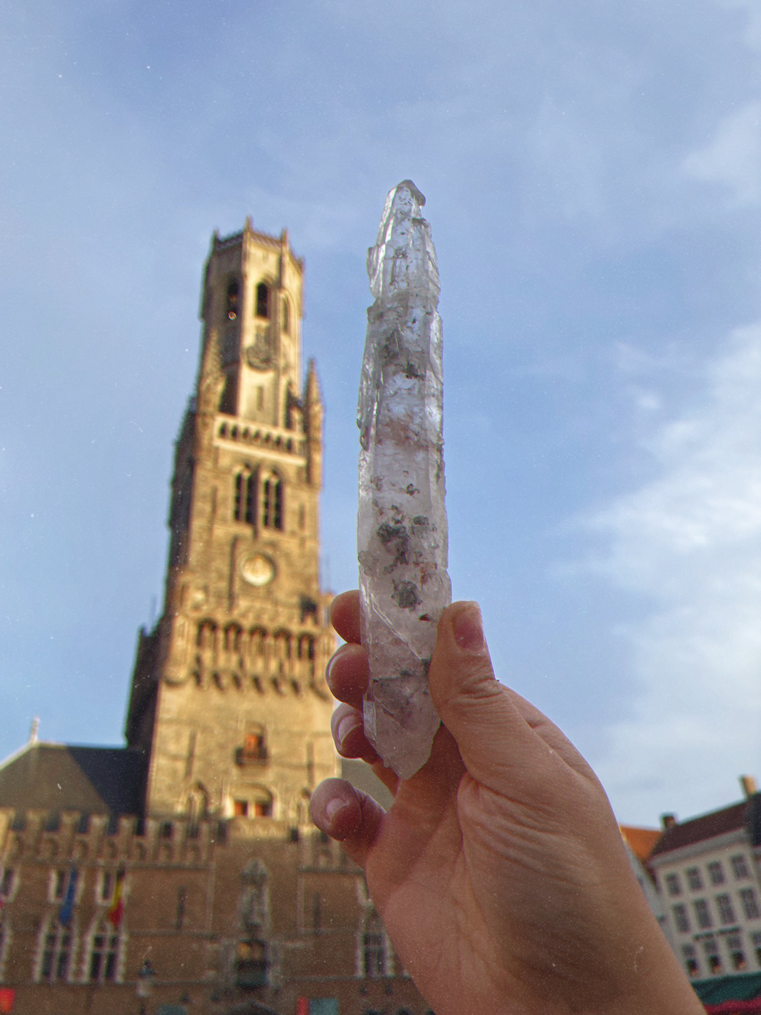 Person holding a large ice stick in front of a historic building and clock tower on a bright, clear day.