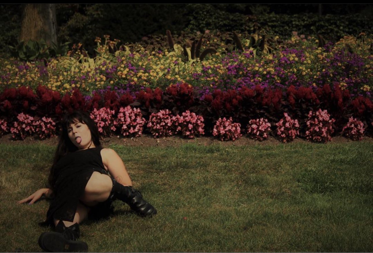 A woman with dark hair, wearing black clothing and boots, lying on the grass with her tongue out, in front of a colorful flower garden at night.