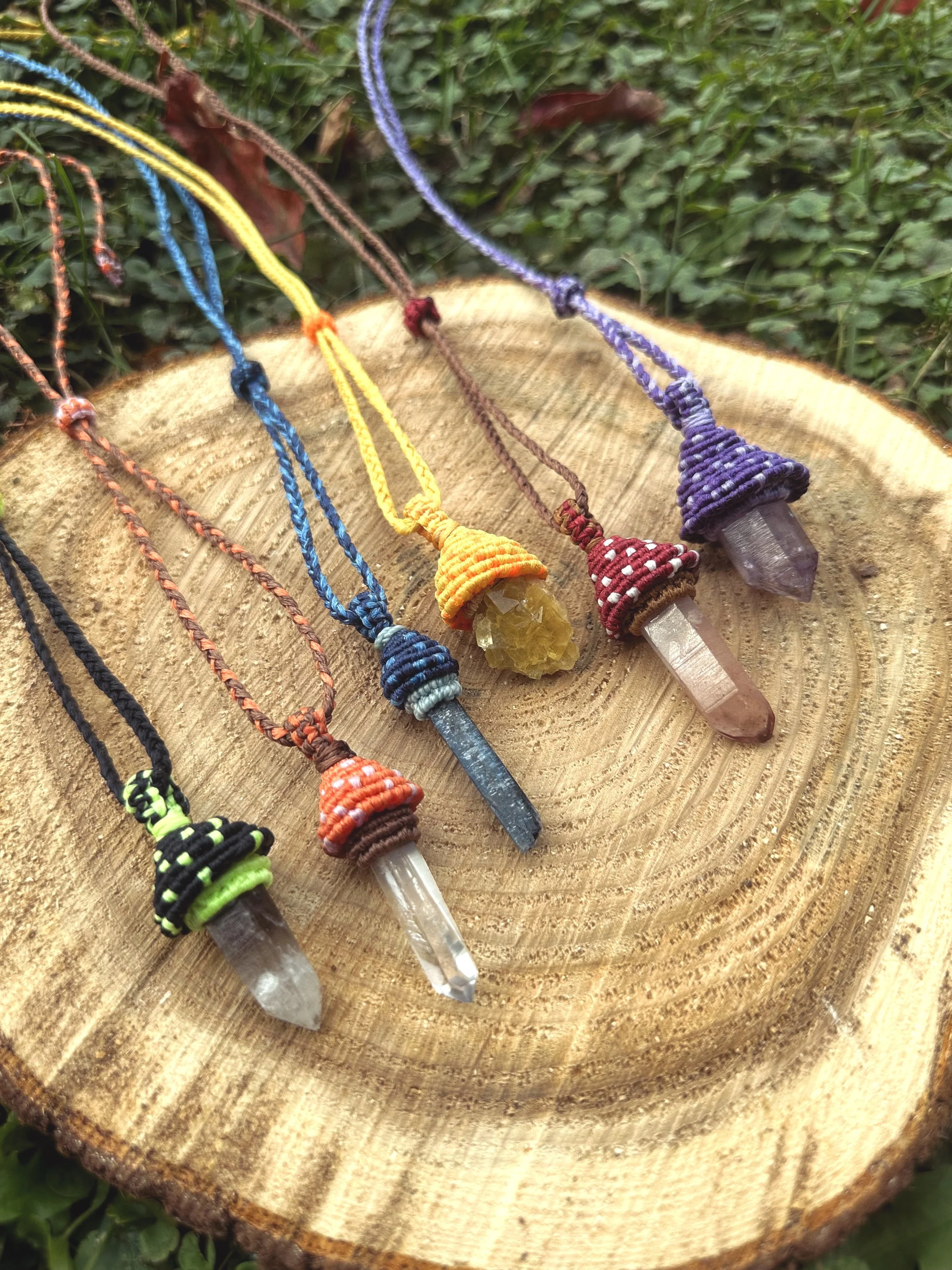Collection of colorful gemstone and crystal pendants with woven cord necklaces, displayed on a wooden surface outdoors.