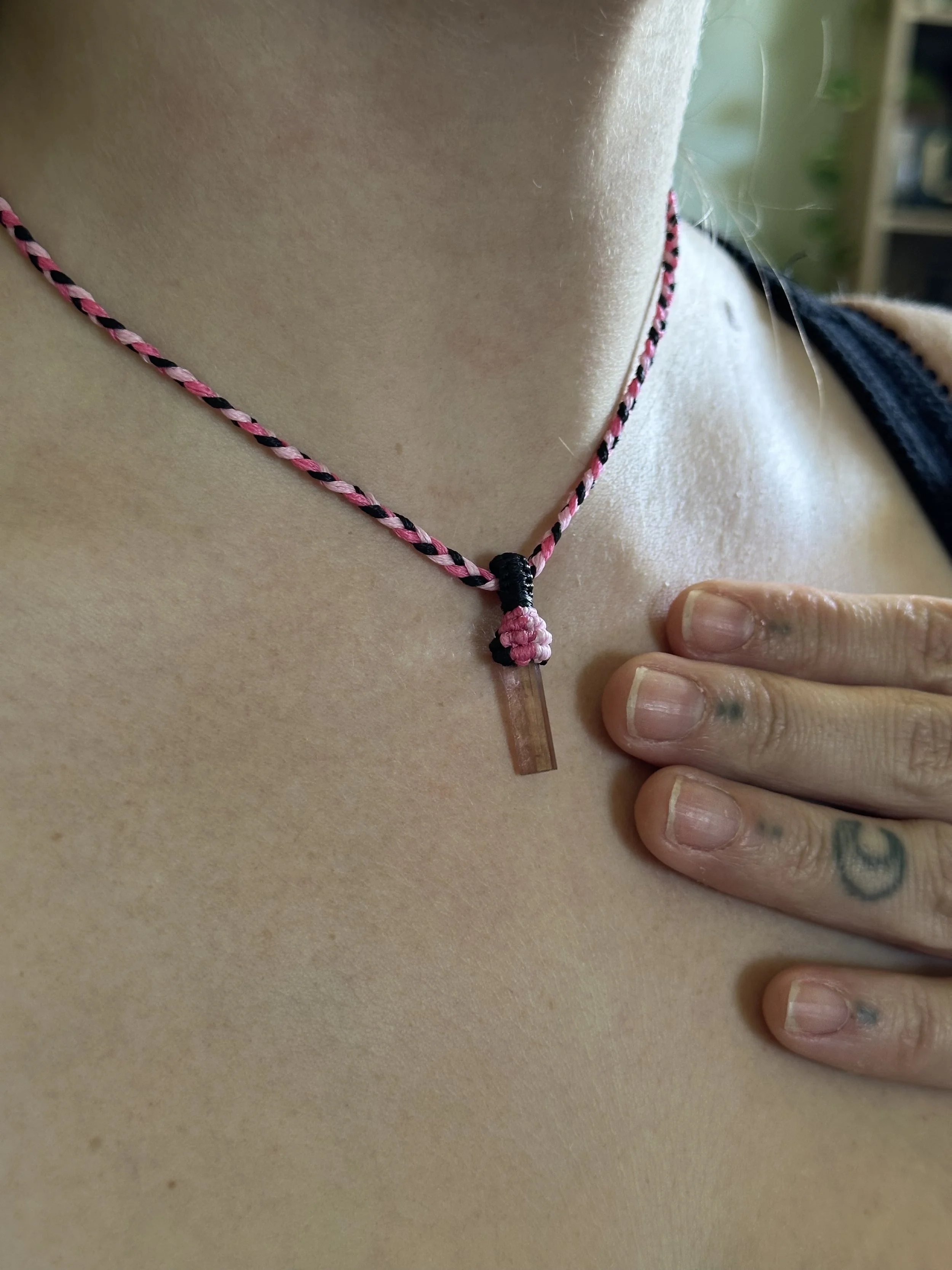 Close-up of a person's neck with a braided necklace and tattooed fingers resting on their collarbone.
