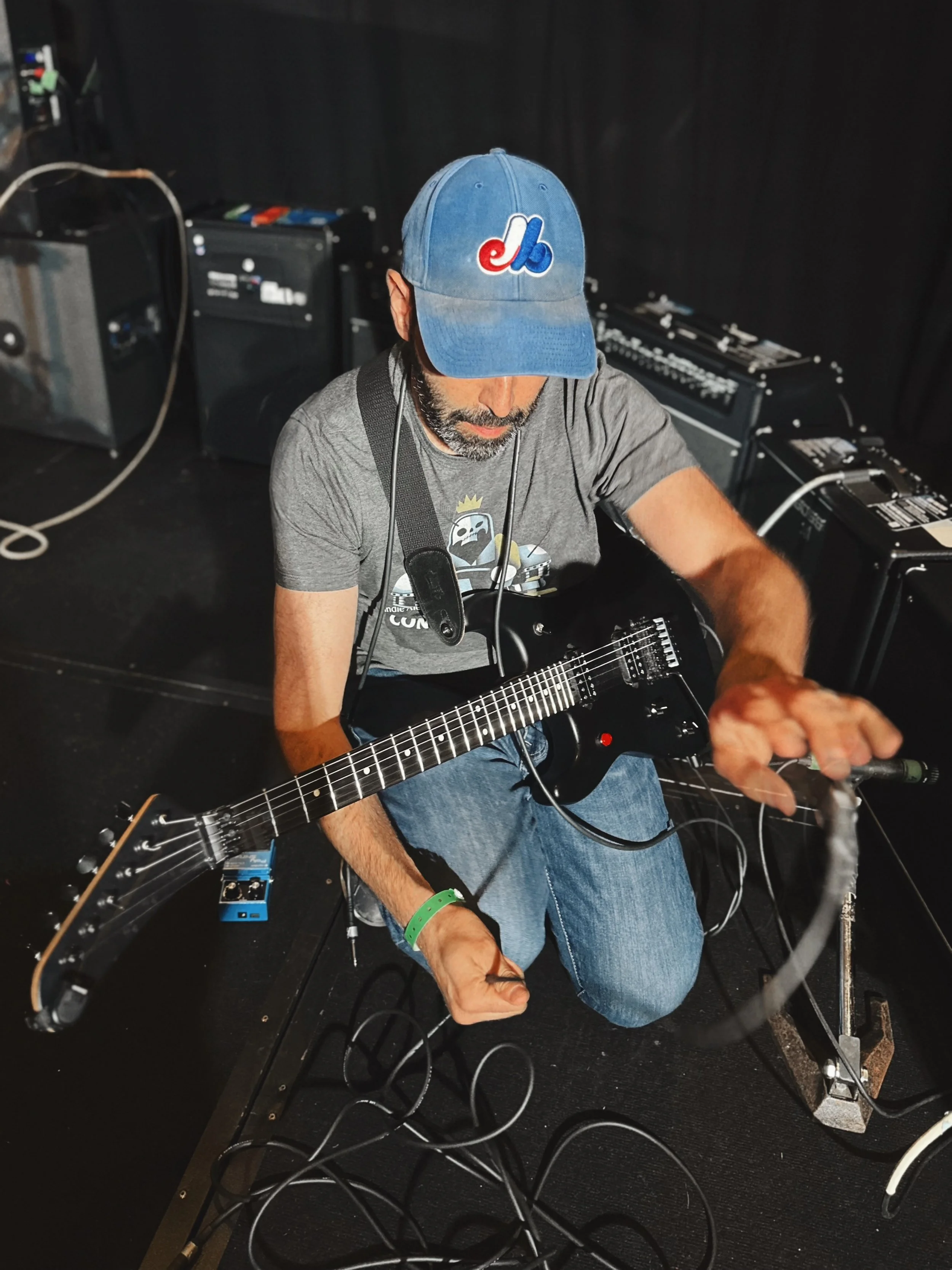 A man sitting on the floor playing an electric guitar, with music equipment and cables around him. He is wearing a blue cap with a logo, a gray t-shirt, and blue jeans.