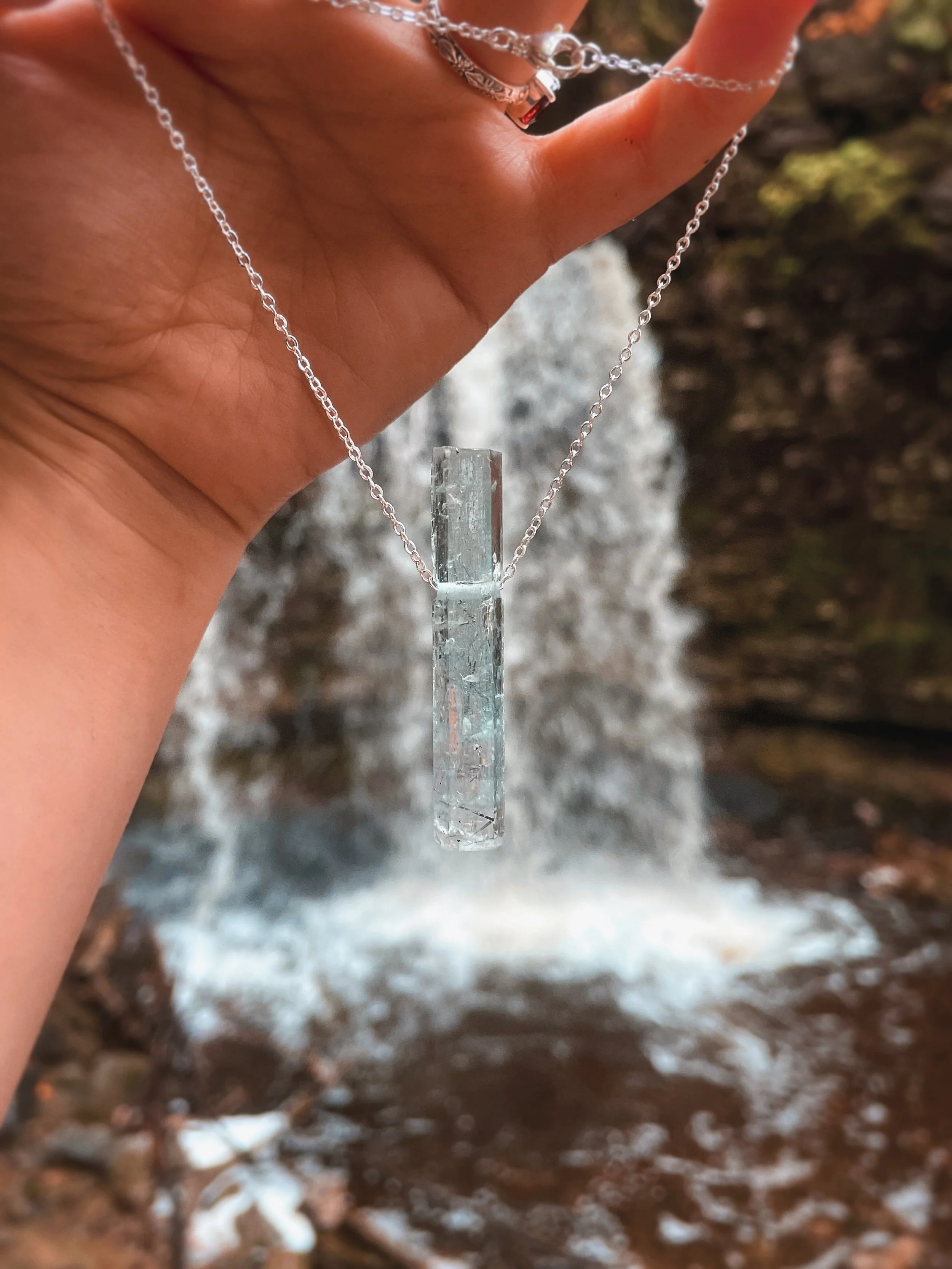 A silver necklace with a transparent rectangular pendant hanging from a person's hand with a background of a waterfall and rocky terrain.