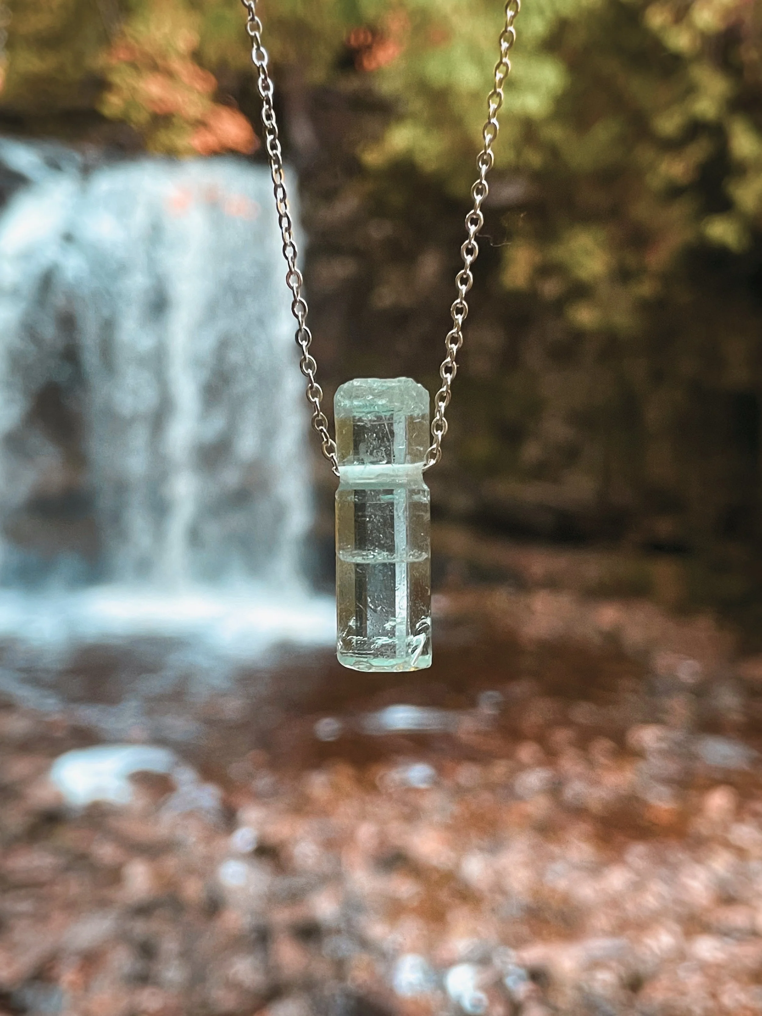A close-up of a delicate necklace with a rectangular, transparent, greenish gemstone pendant hanging from a silver chain, with a blurred outdoor background featuring a waterfall and autumn foliage.