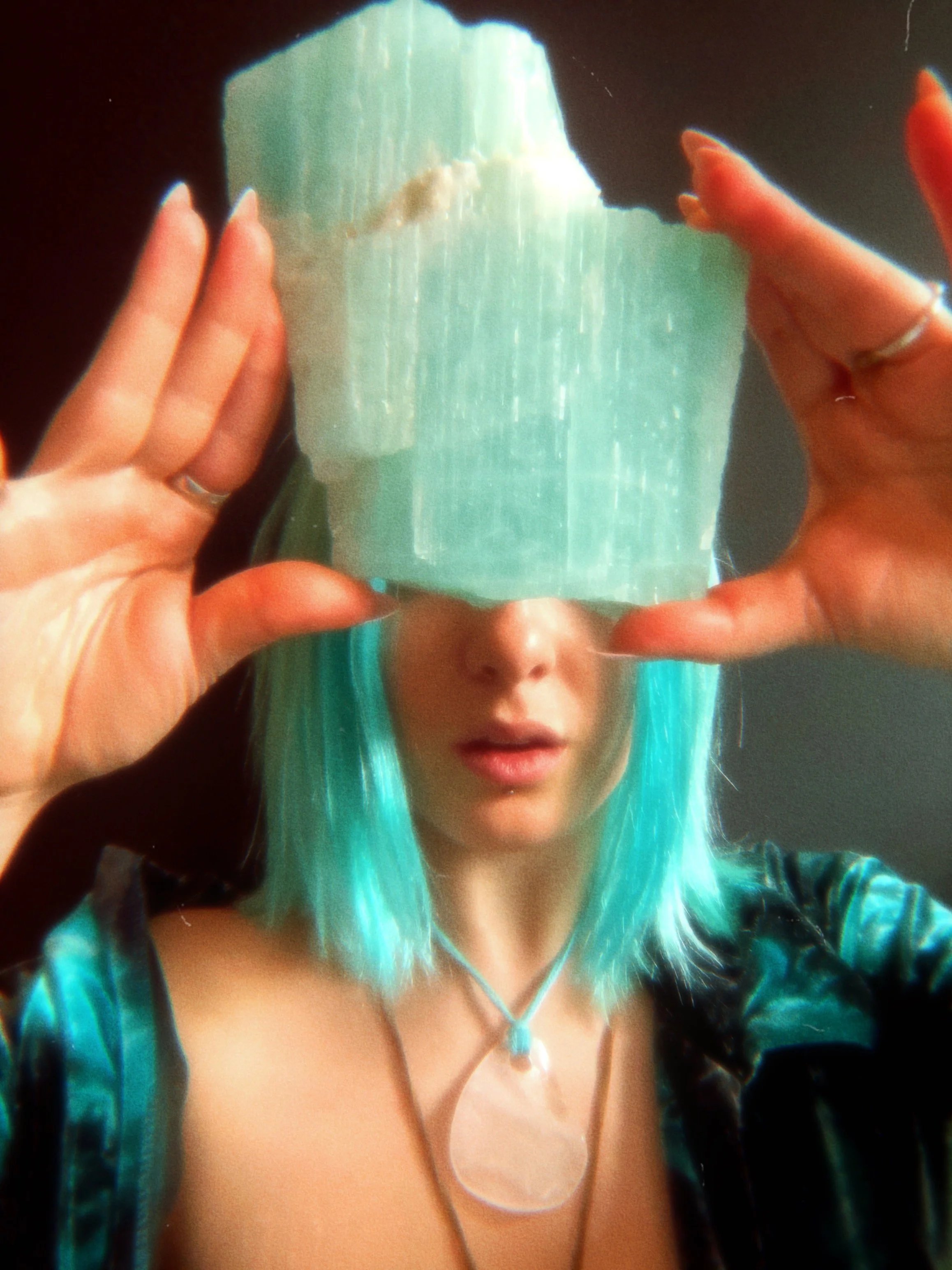 A person with teal hair and a black leather jacket taking a selfie through a shiny reflective surface, showing their face partially, with a focus on their hair and jewelry.
