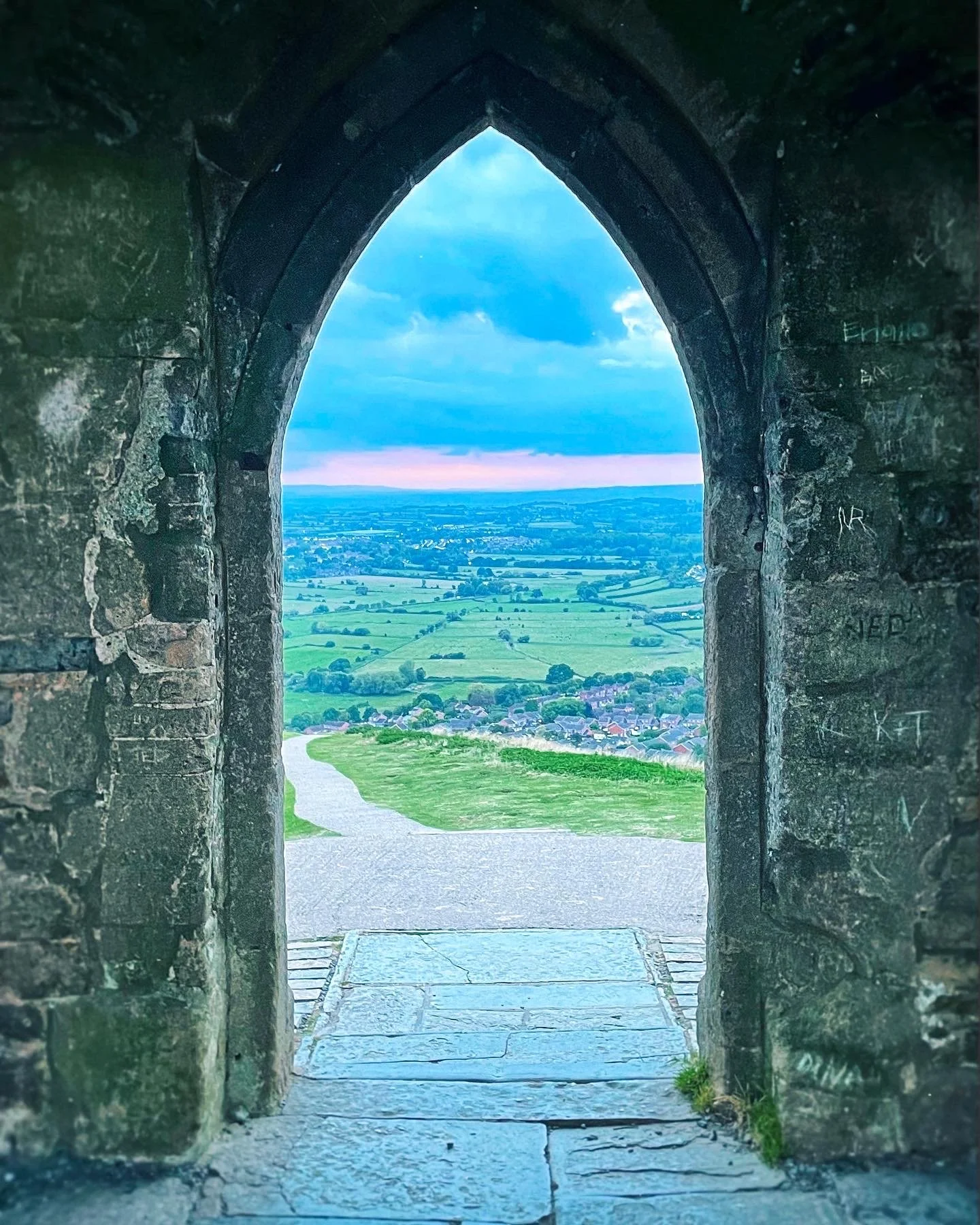 View through a stone archway of a rural landscape with fields, trees, and a small town or village under a partly cloudy sky.