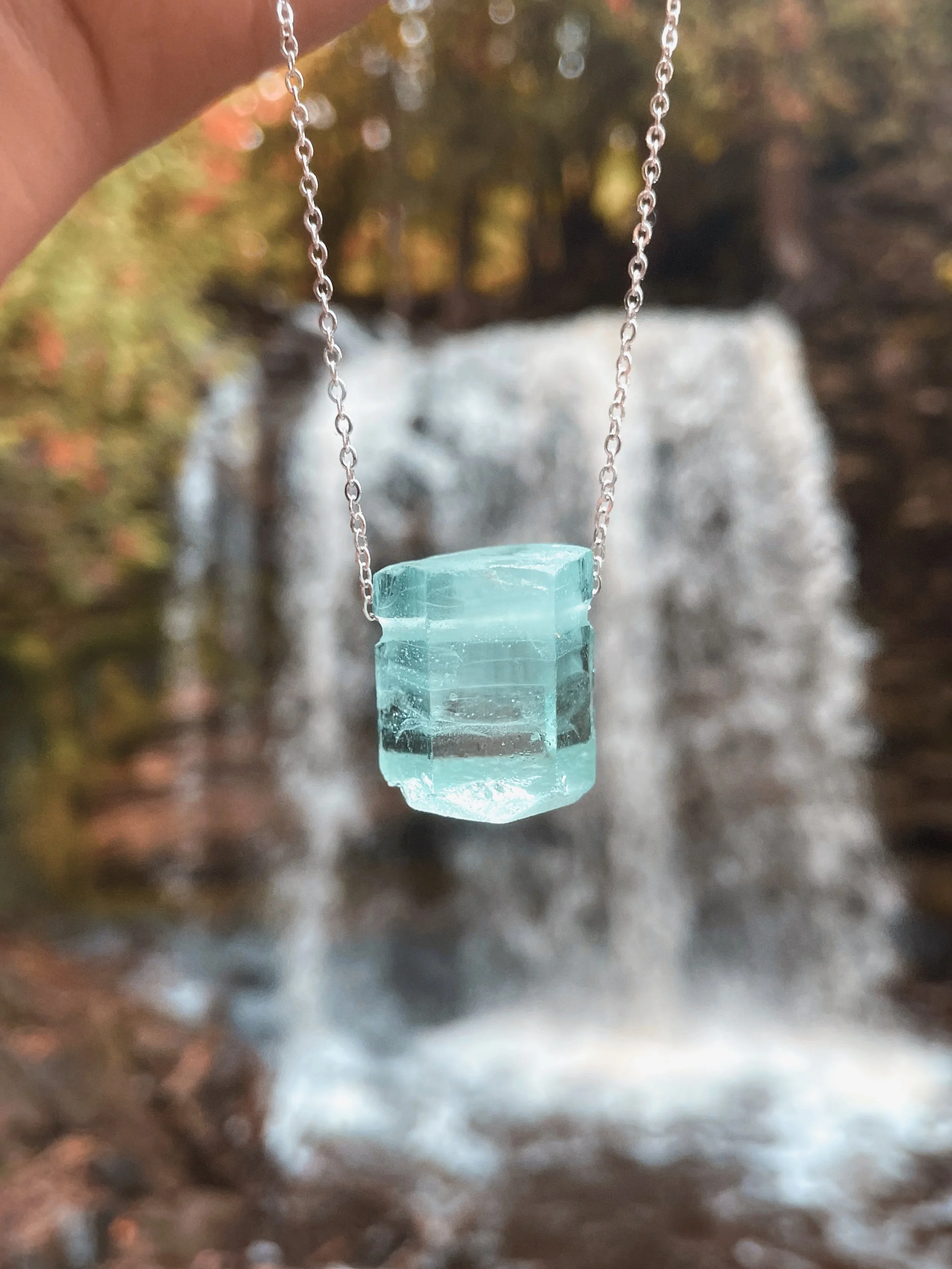 A close-up of a silver chain necklace with a turquoise glass or gemstone pendant hanging against a blurred outdoor background with trees and rocks.