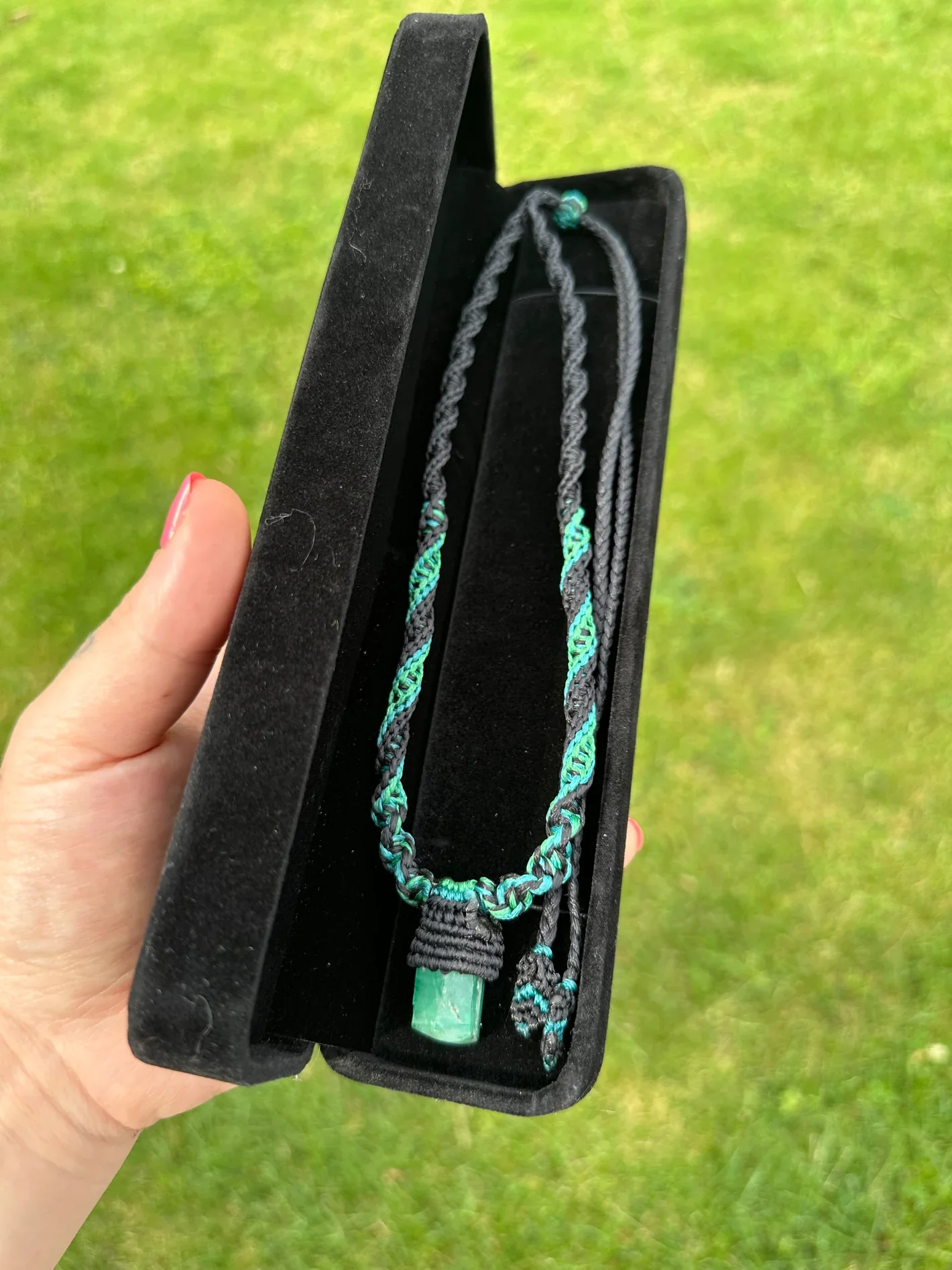 A hand holding an open black jewelry box containing a braided necklace with green and black strands and a green pendant, set against a grassy background.