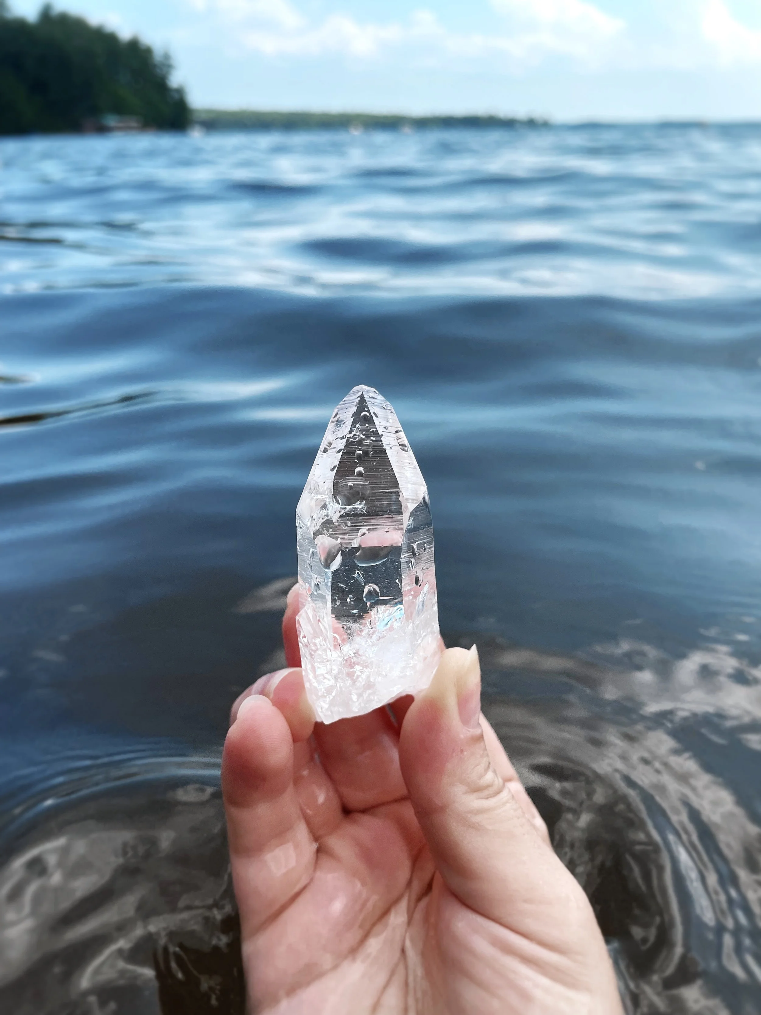 A person holding a clear quartz crystal at the edge of a body of water with a coastline and cloudy sky in the background.