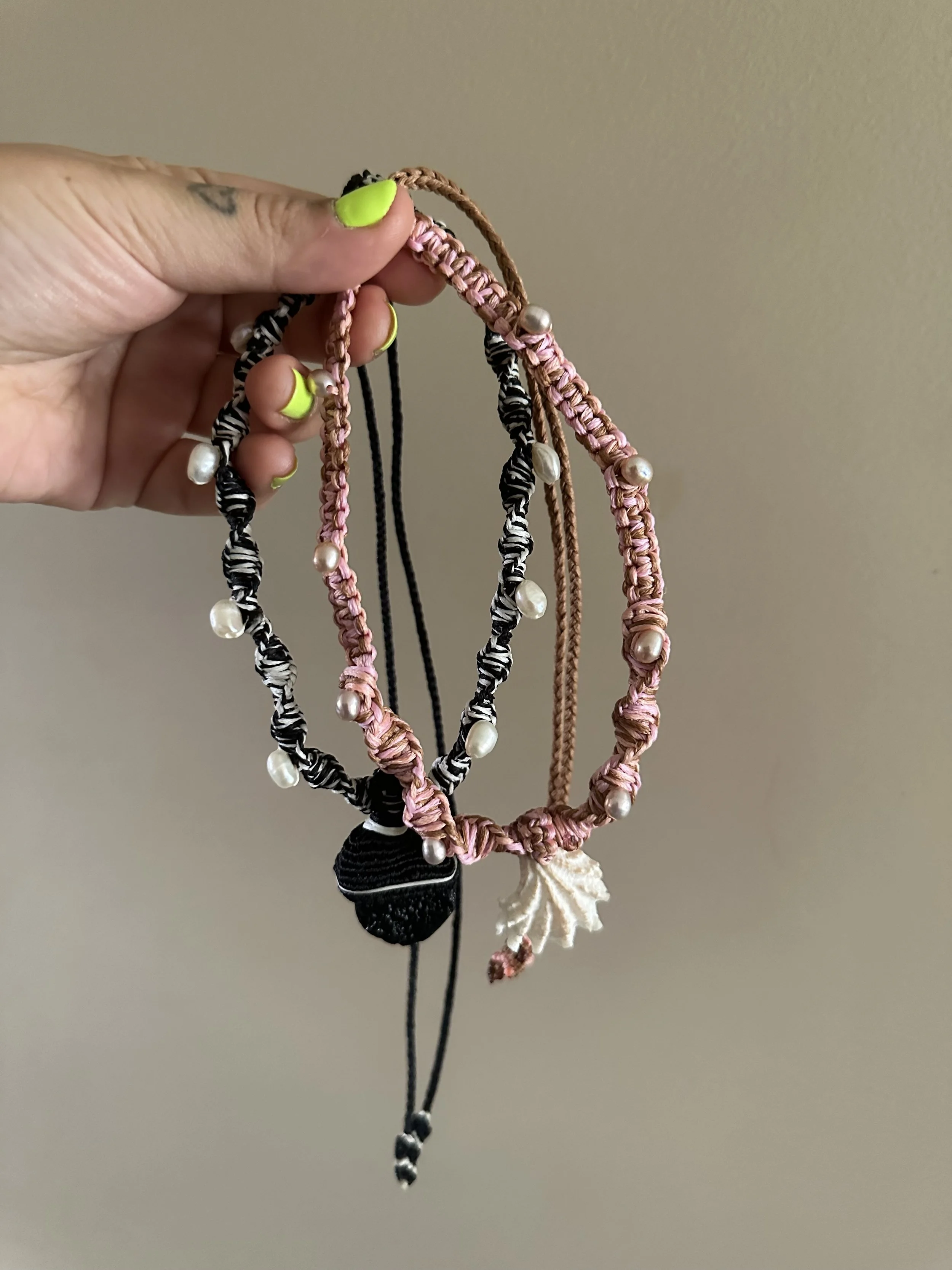 Hand holding four woven, beaded friendship bracelets made of pink and brown cords, with pearls and tassels, against a plain gray background.