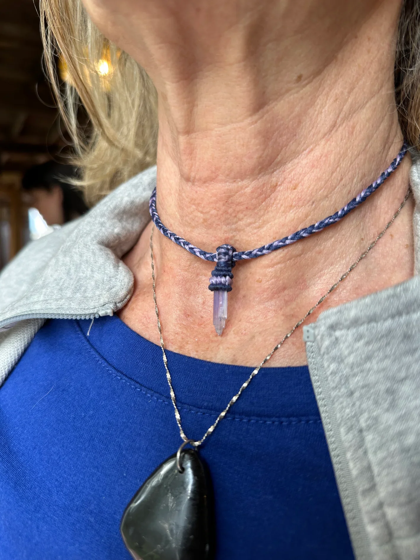 Close-up of a person's neck wearing a purple braided necklace with a pointy crystal pendant, layered with a thin silver chain and a black stone pendant necklace, with a gray hoodie and blue shirt. Part of a person's face and hair and another person b