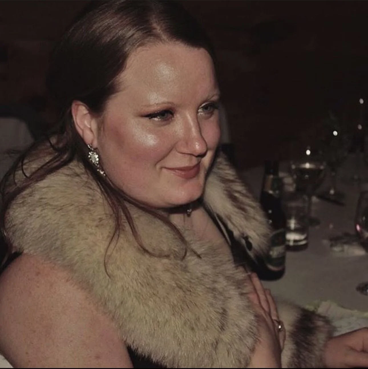 A woman with long brown hair, wearing earrings and a fur stole, smiling at a dinner event with glasses and bottles on the table.