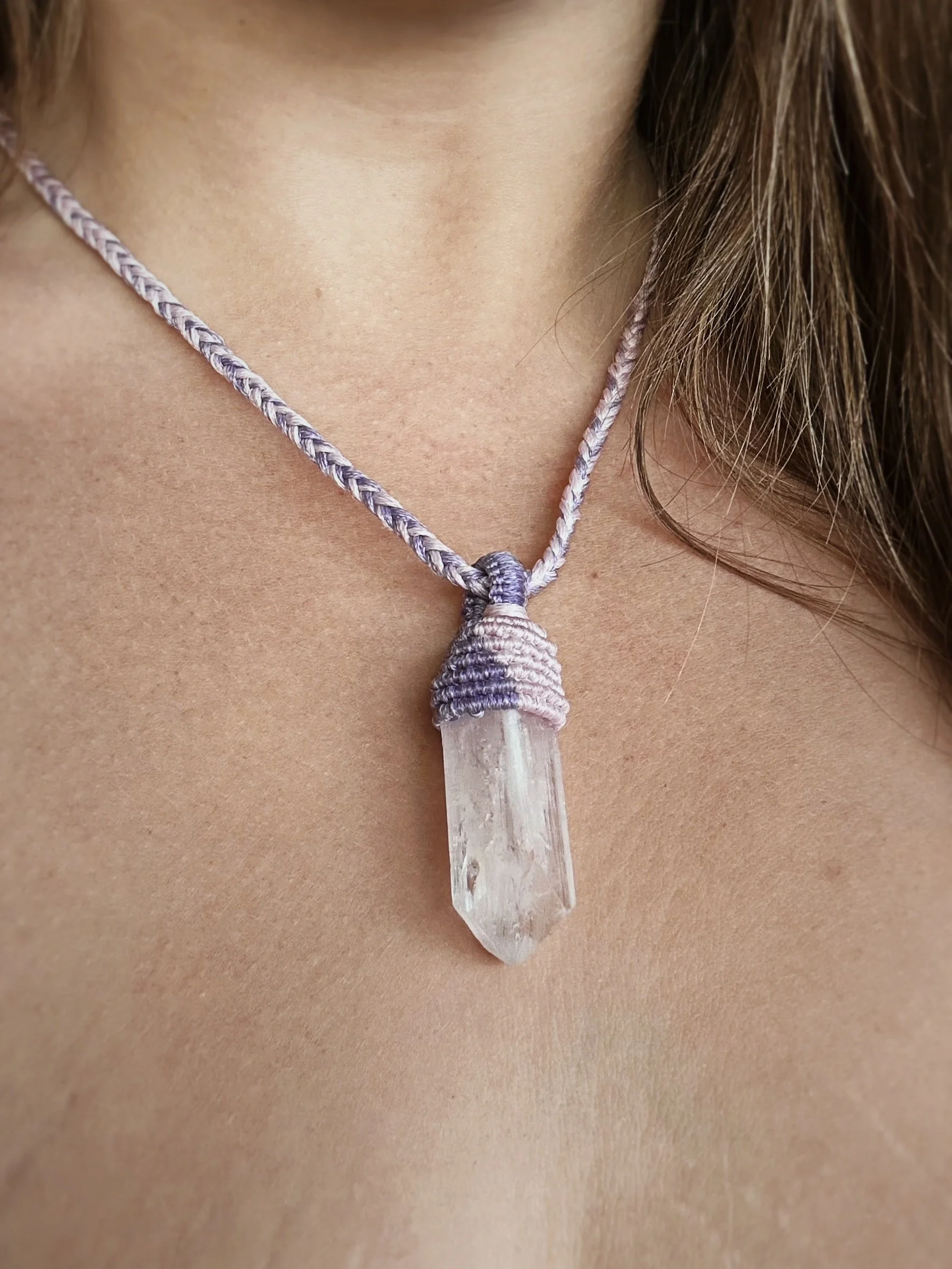 A close-up of a person wearing a necklace with a clear quartz crystal pendant tied with purple and white braided cord.