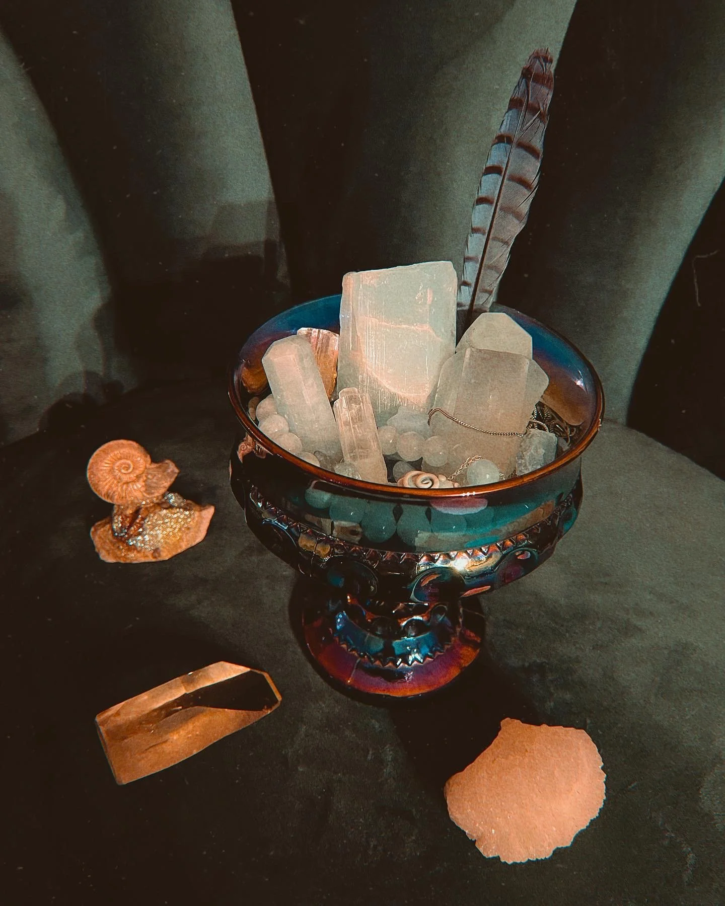 A decorative glass bowl with large clear quartz stones and feathers, placed on a dark surface with a shell and a piece of amber-colored stone nearby.