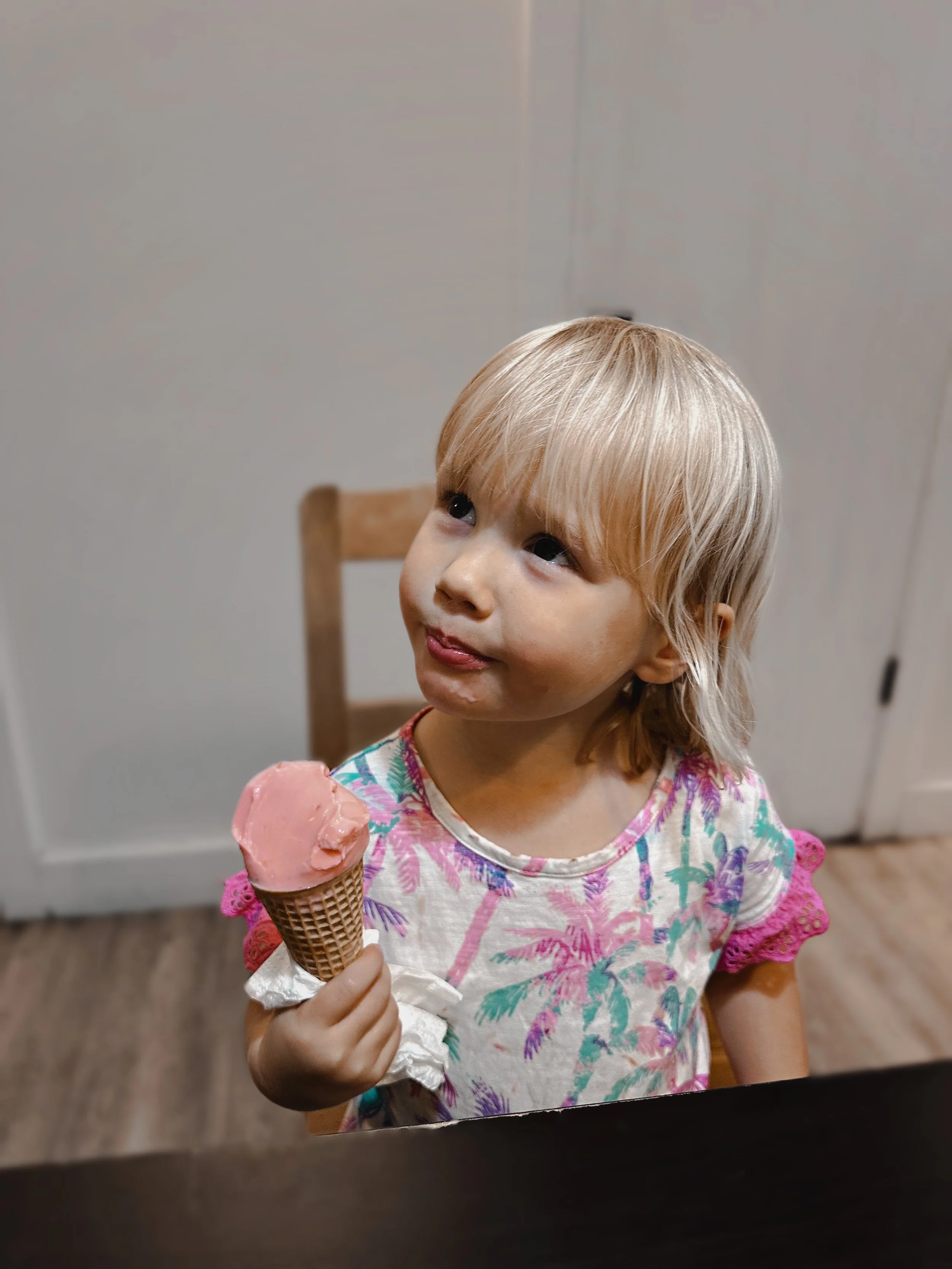 A young girl with blonde hair holds a pink ice cream cone and looks up with a smile.