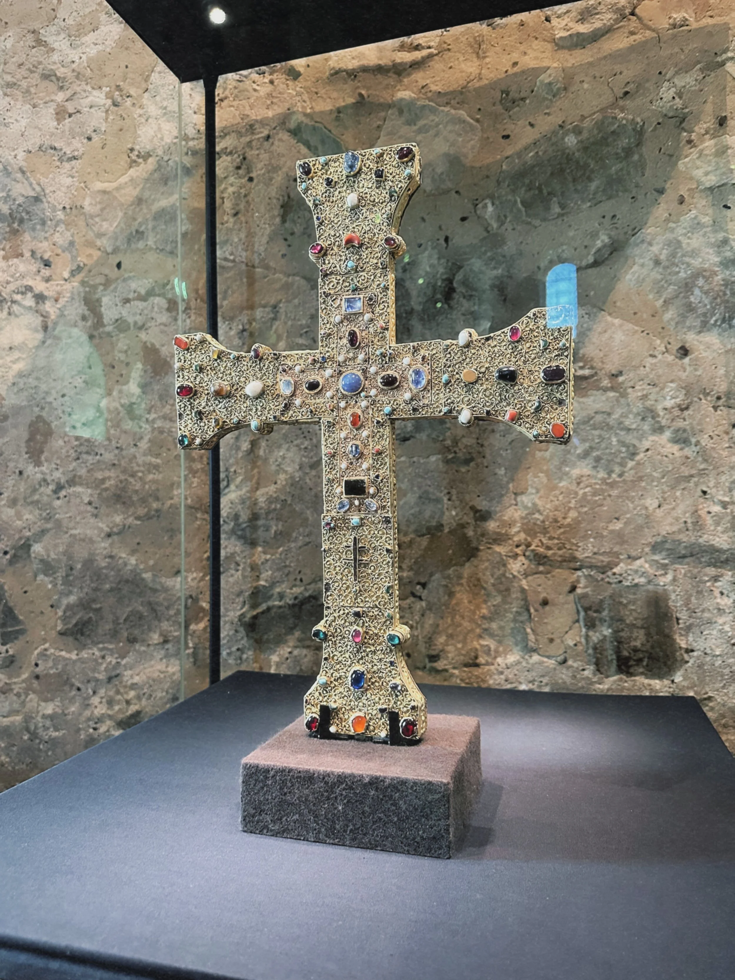 An ornate gold cross adorned with numerous colorful gemstones is displayed inside a glass case on a stand in a museum or exhibition setting with a rocky wall background.