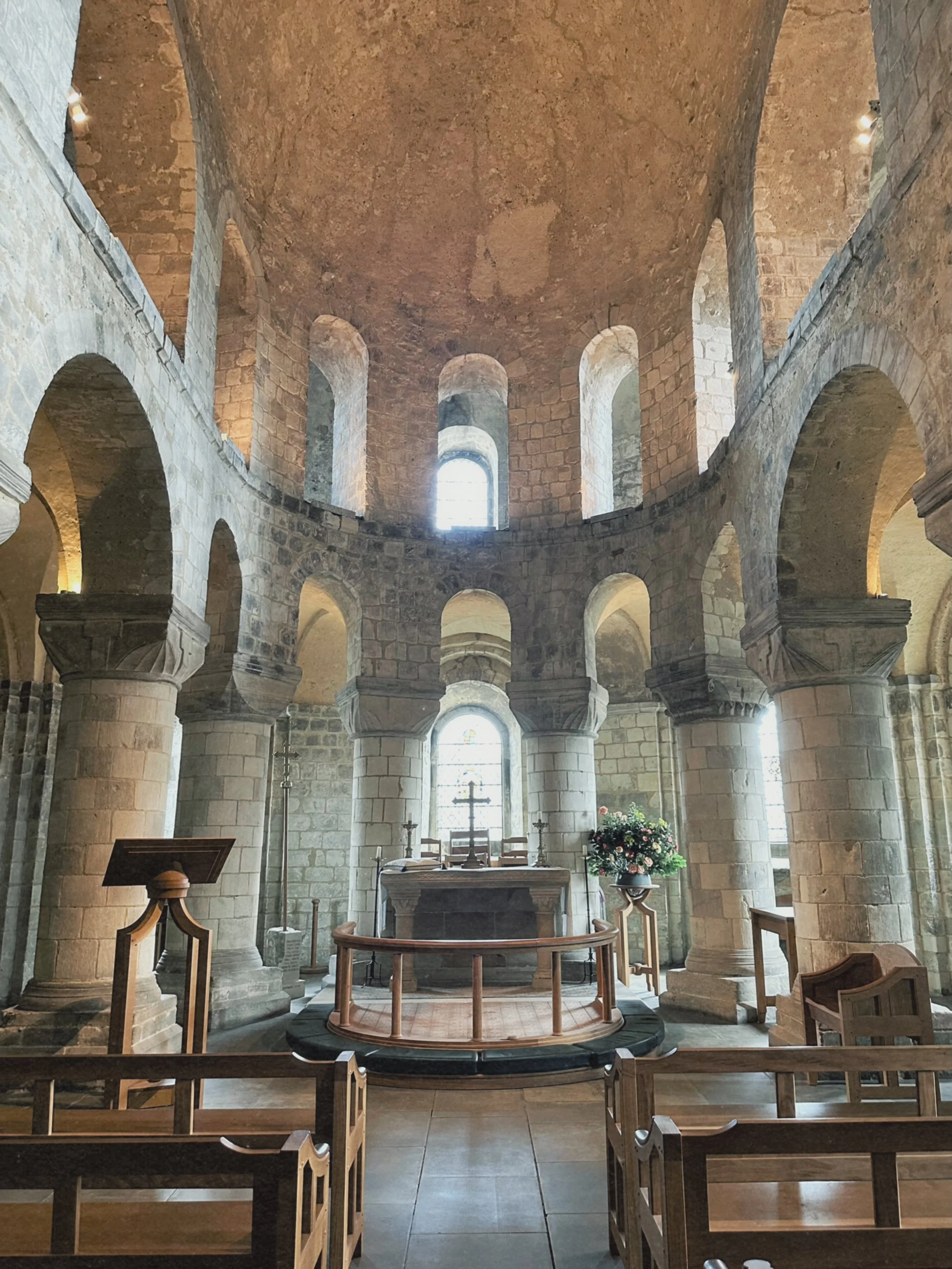 Interior of a historic stone church with an altar, surrounded by wooden pews, columns, and arched windows, with a cross and floral arrangement at the altar.