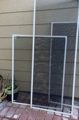 Two screen doors leaning against the house wall next to potted plants on a brick patio.