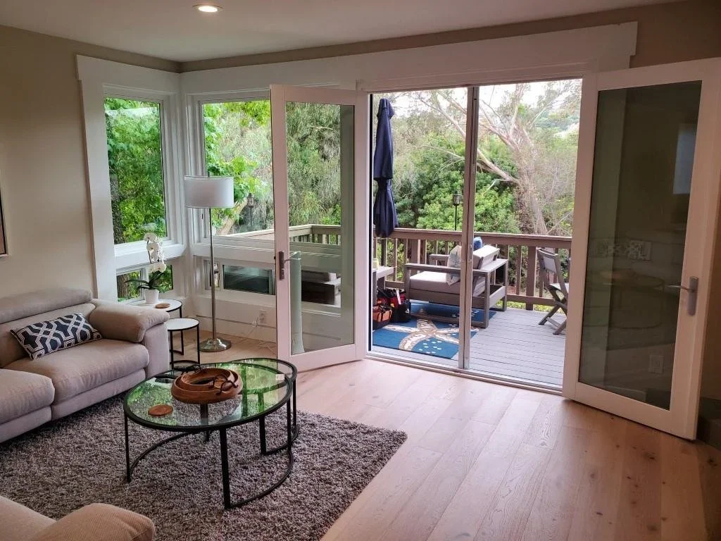 Living room with open sliding glass door leading to a wooden deck with outdoor furniture and trees outside.