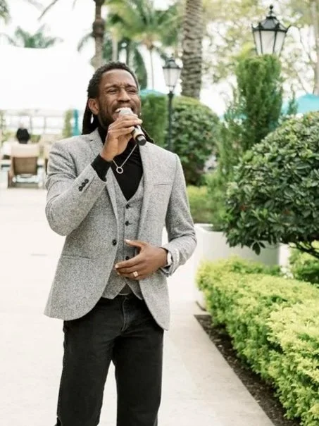 Man in gray blazer singing into a microphone outdoors with greenery and street lamps in the background.
