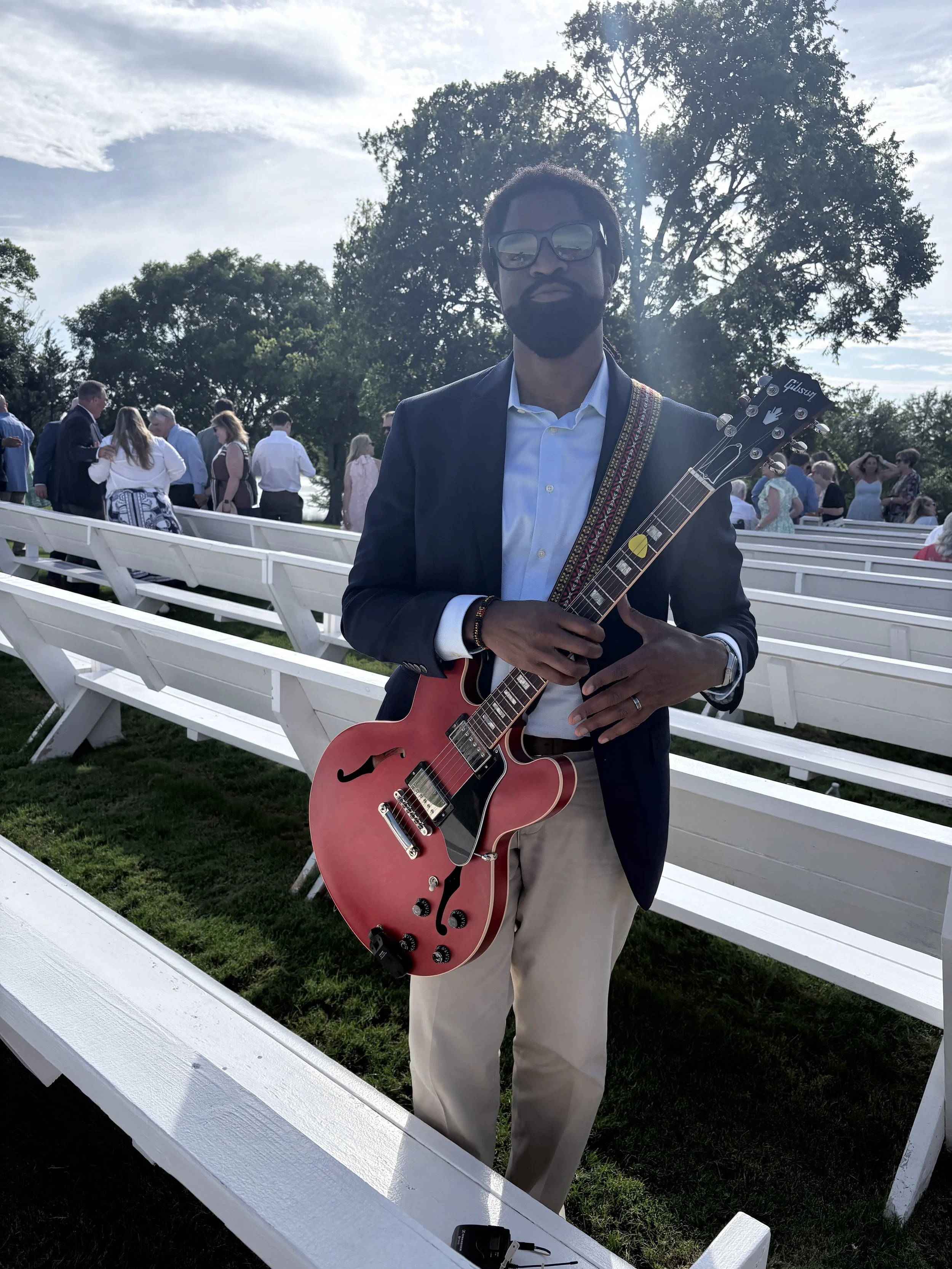 Man in a suit jacket and sunglasses holding a red electric guitar at an outdoor event with people standing and chatting in the background, white benches and trees under a partly cloudy sky.