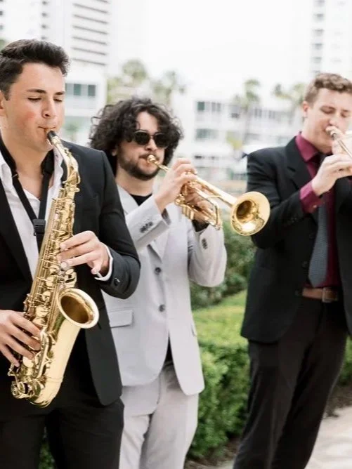 Three men in suits playing saxophone, trumpet, and clarinet outdoors in an urban setting.