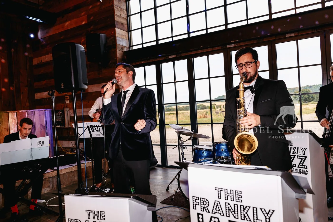 Jazz band performing indoors with large window view, featuring a singer and a saxophonist, with a pianist and drummer in the background, at an event with a sign reading "The Frankly Jazz Band."