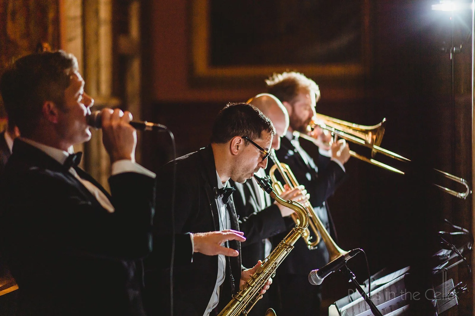 Jazz band performing with three musicians playing saxophones and one on trumpet, dressed in black tuxedos, lights focused on them in a dimly lit room.