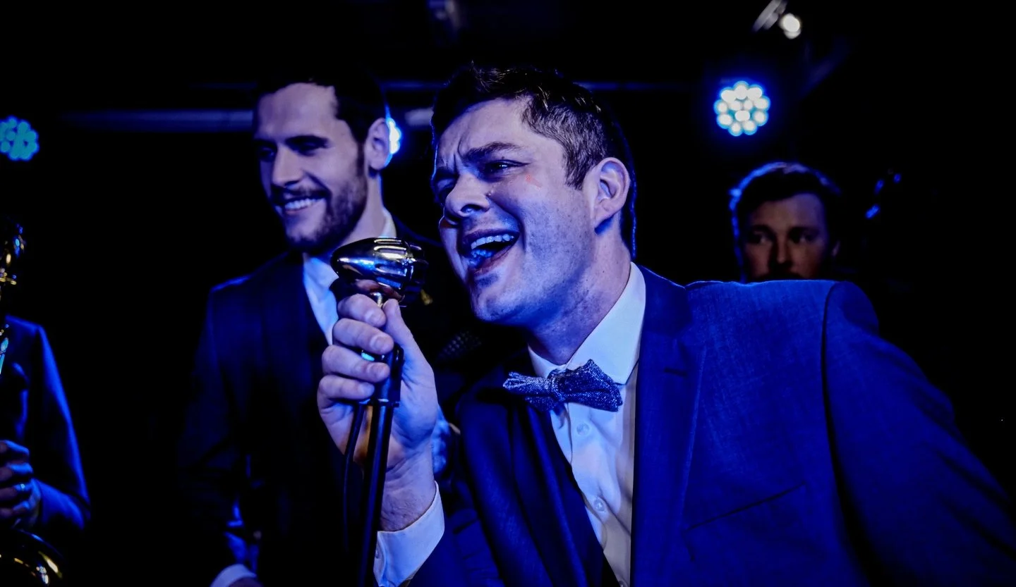 A man singing into a microphone with a joyful expression in a dark setting with blue lights, surrounded by other people, some of whom are smiling.