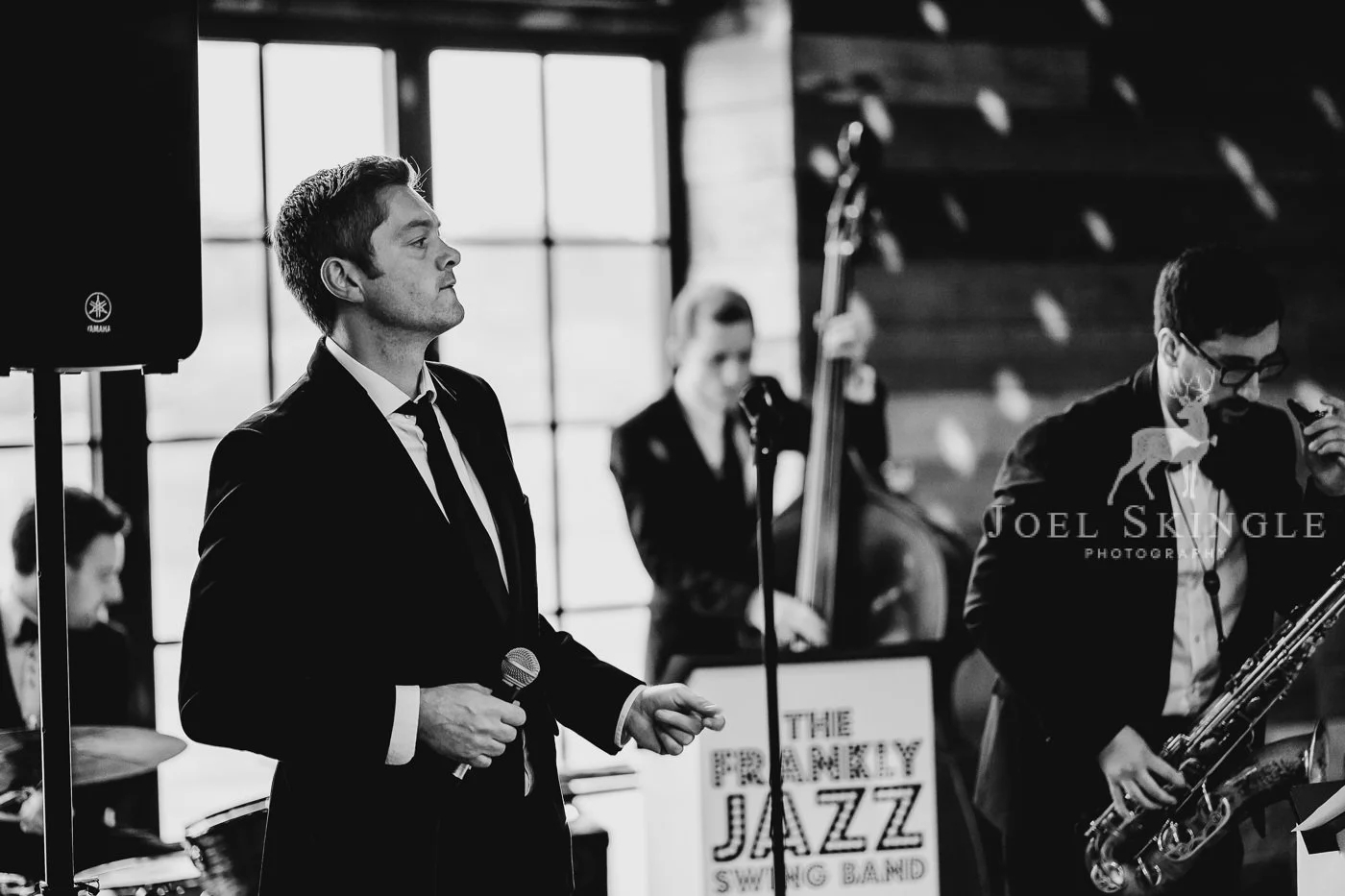 Black and white photo of a jazz band performing live, with a man in a suit holding a microphone in the foreground, and other band members playing instruments in the background, including a double bass and a saxophone.