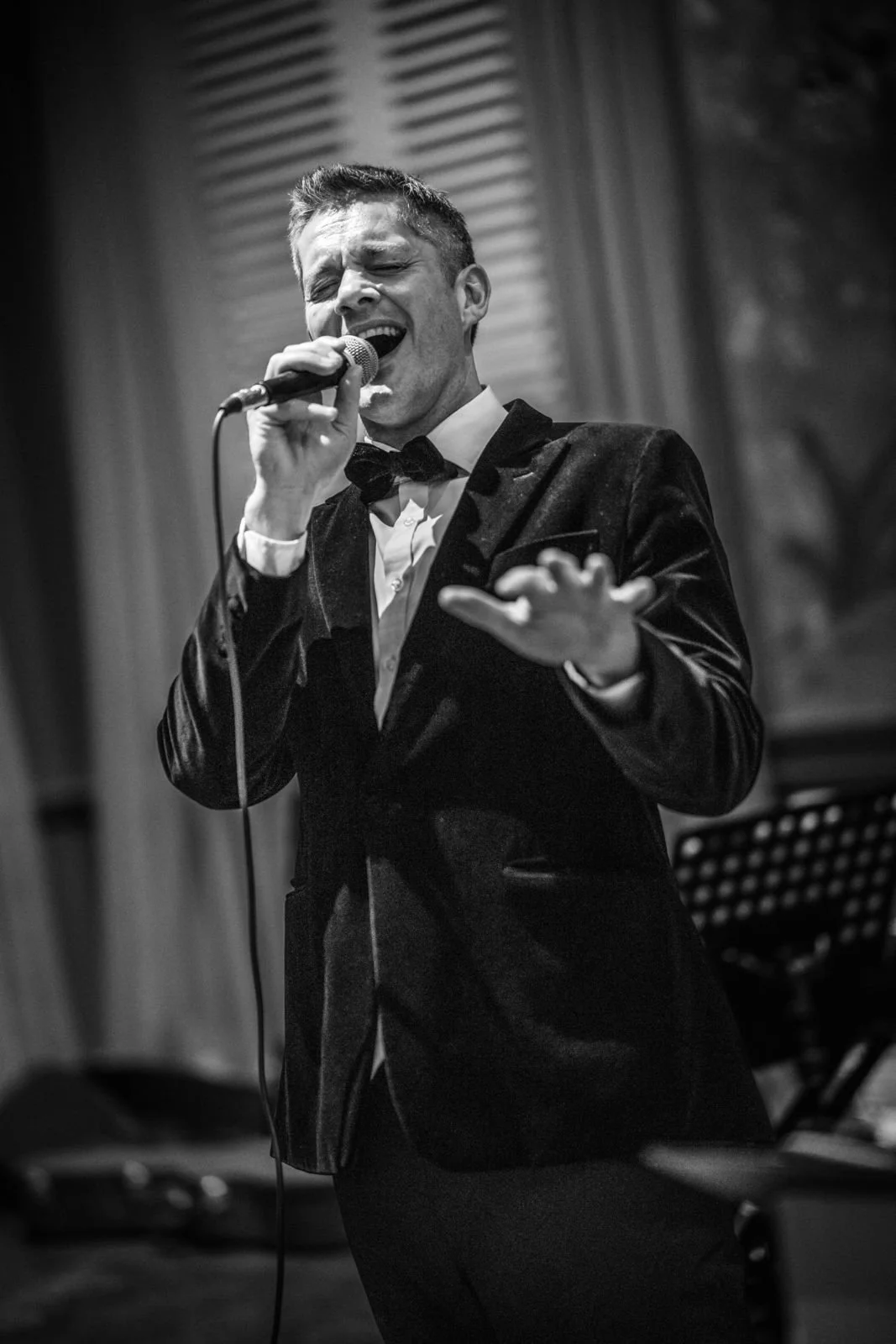 A man in a tuxedo singing into a microphone with a hand raised, performing passionately on stage.