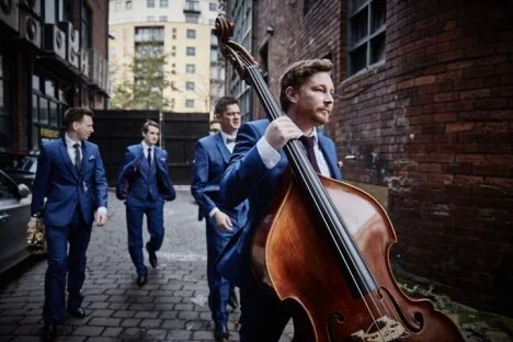 A man playing a double bass outdoors with three men in suits walking behind him in an alleyway.