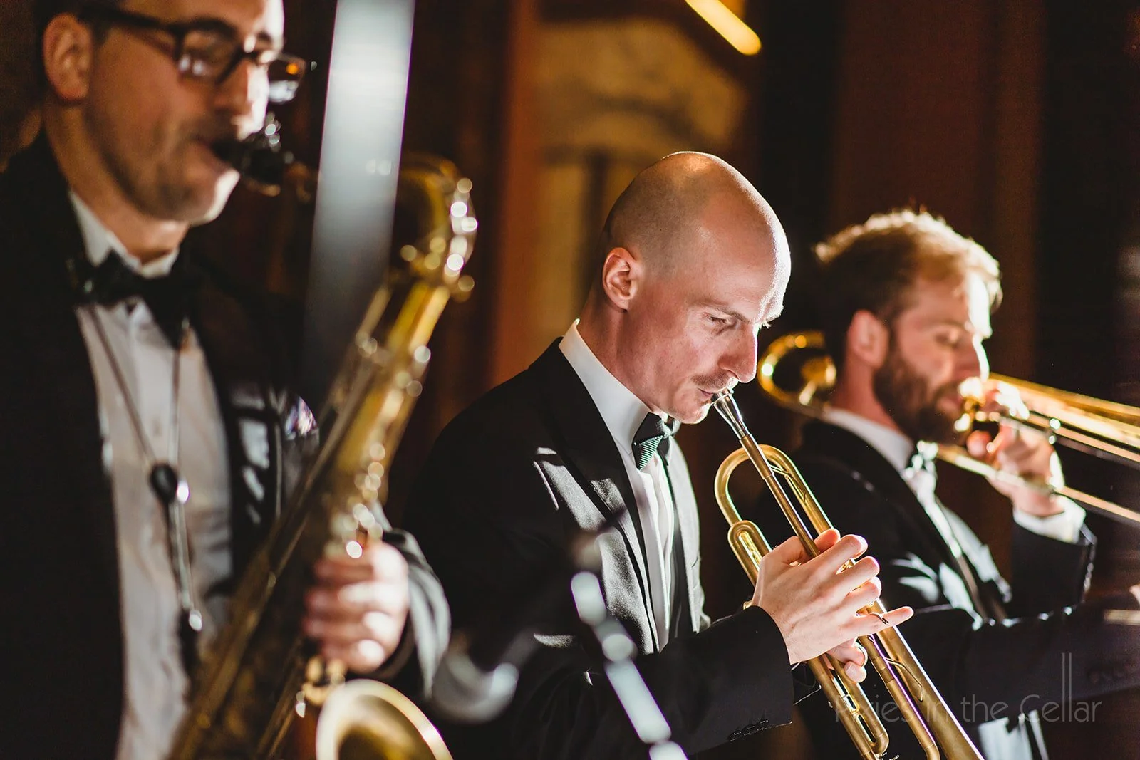Three jazz musicians in tuxedos playing wind instruments in a dimly lit setting.