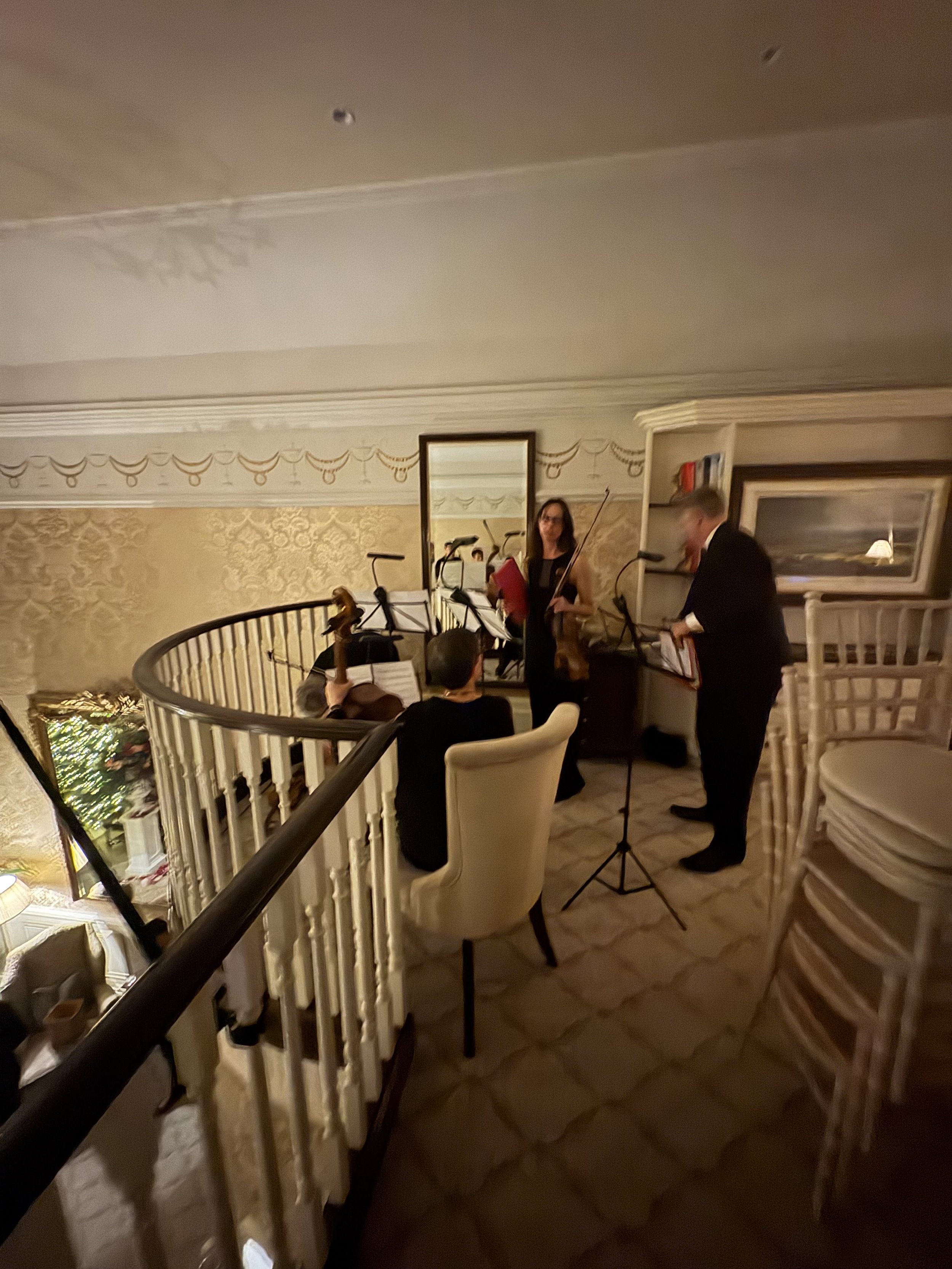 A woman and man playing violins at a music event in an elegant, warmly lit room with beige walls, decorative molding, a large mirror, and potted plants. The woman is standing near a music stand, and the man is standing on the right side.