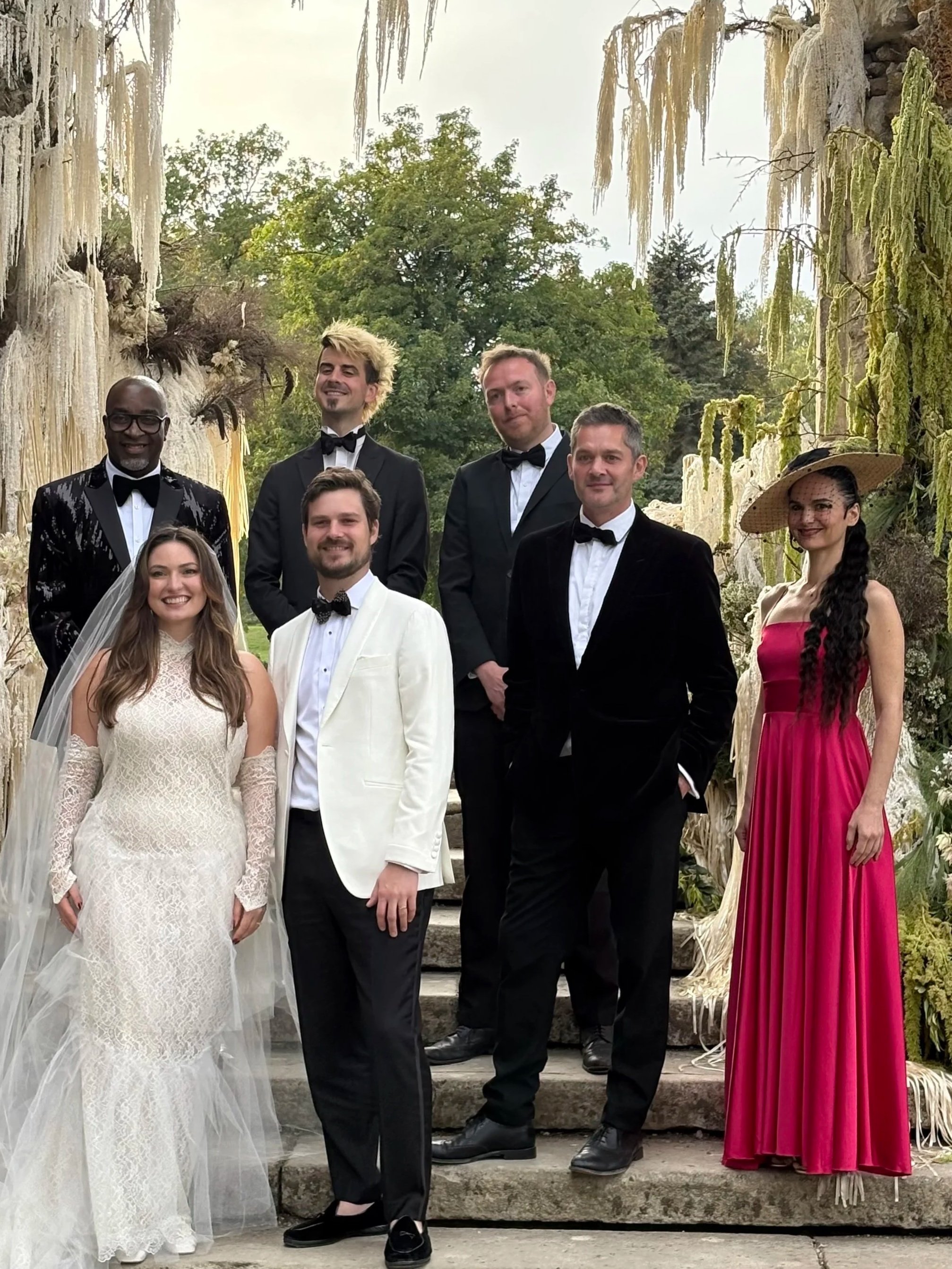 A group of seven people dressed in formal and vintage attire standing on stone steps outdoors, with trees and hanging moss in the background.