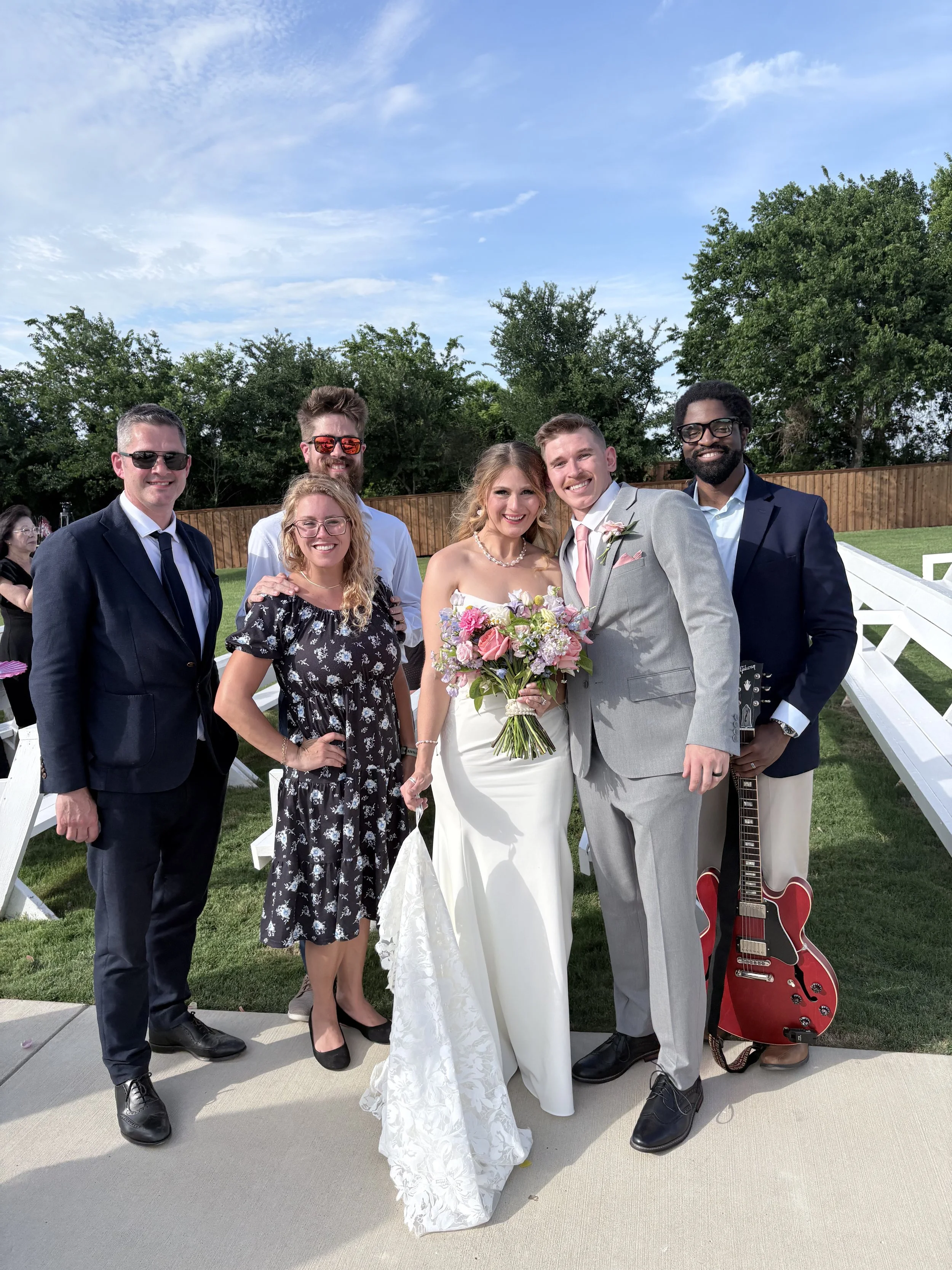 A group of seven people posing together outdoors, dressed in formal attire, with two women in wedding dresses and the others in suits. The background features trees, a wooden fence, and a partly cloudy sky.