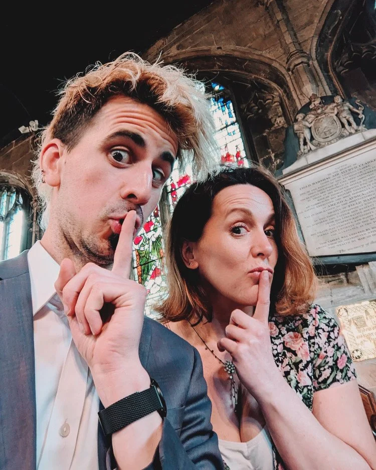 A man and woman inside a church, both holding a finger to their lips in a 'shh' gesture, with stained glass windows and religious plaques in the background.